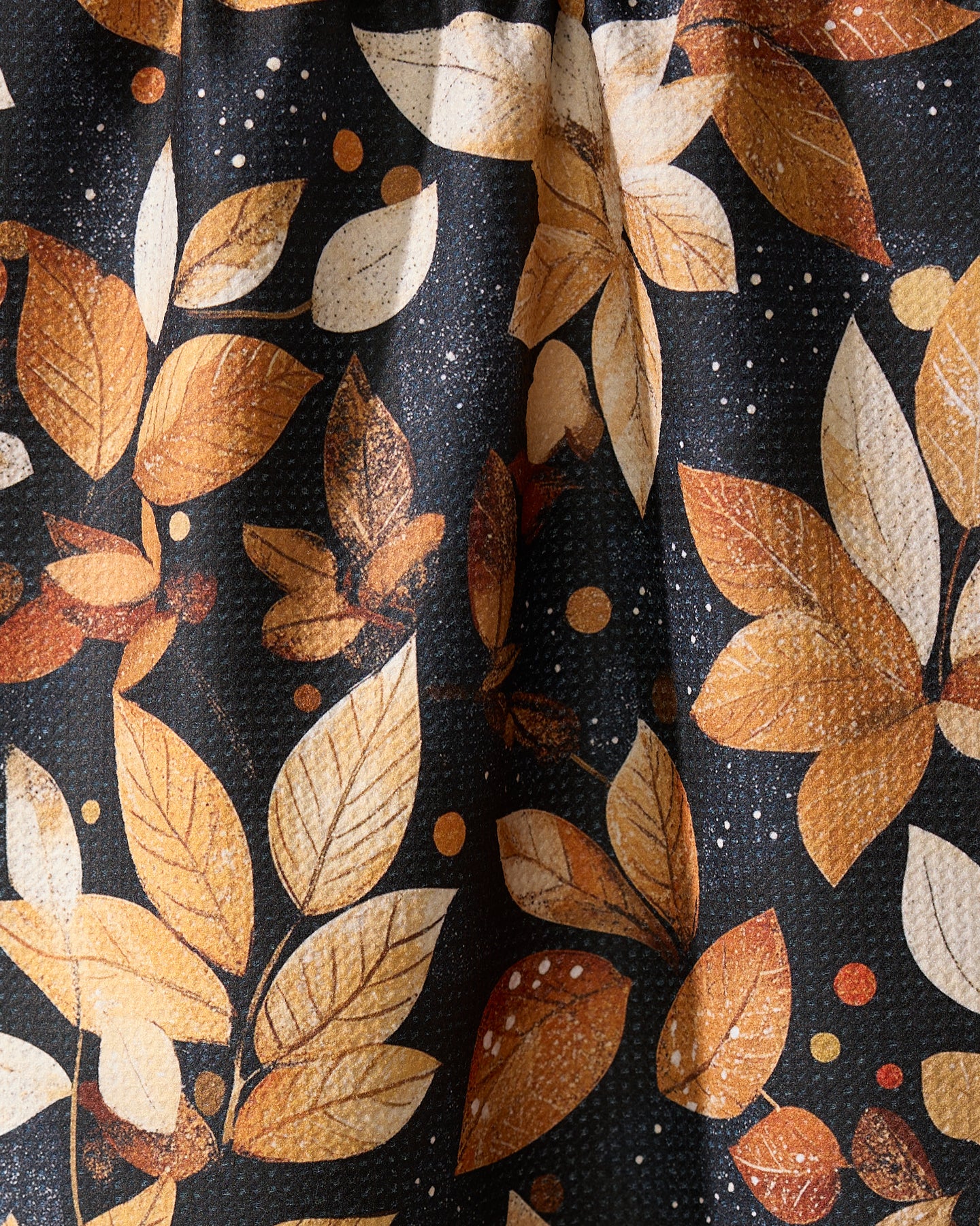 A close-up of Geometry’s Botanical Wonderland towel shows orange, yellow, and white autumn leaves with small dots on a dark background. Ultra-absorbent with visible texture and folds, it’s perfect as a kitchen tea towel.
