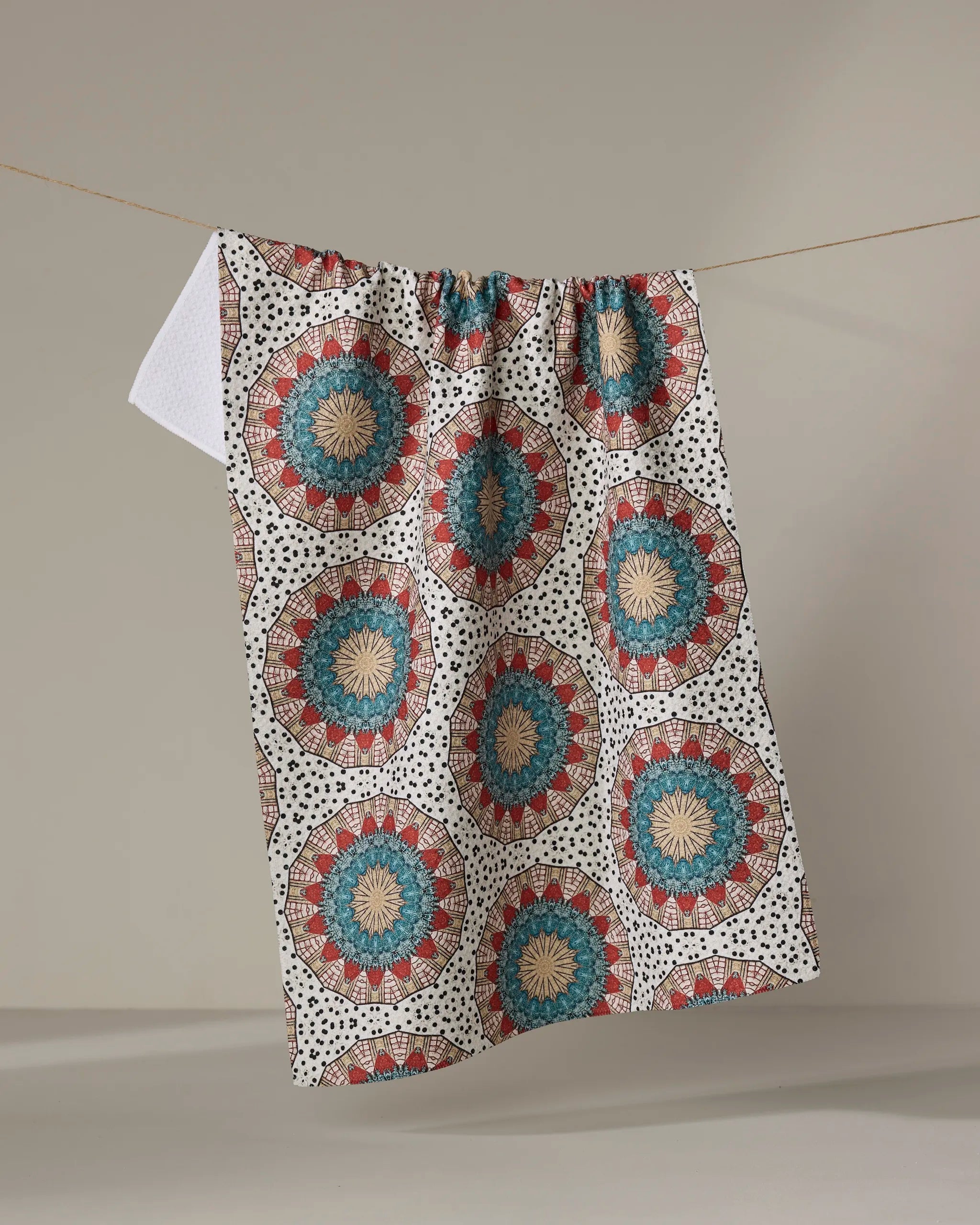 The Katherine Berkeley "Bottle Cap Boogie Woogie" tea towel, with circular blue, red, and beige designs, hangs from a string against a plain background.