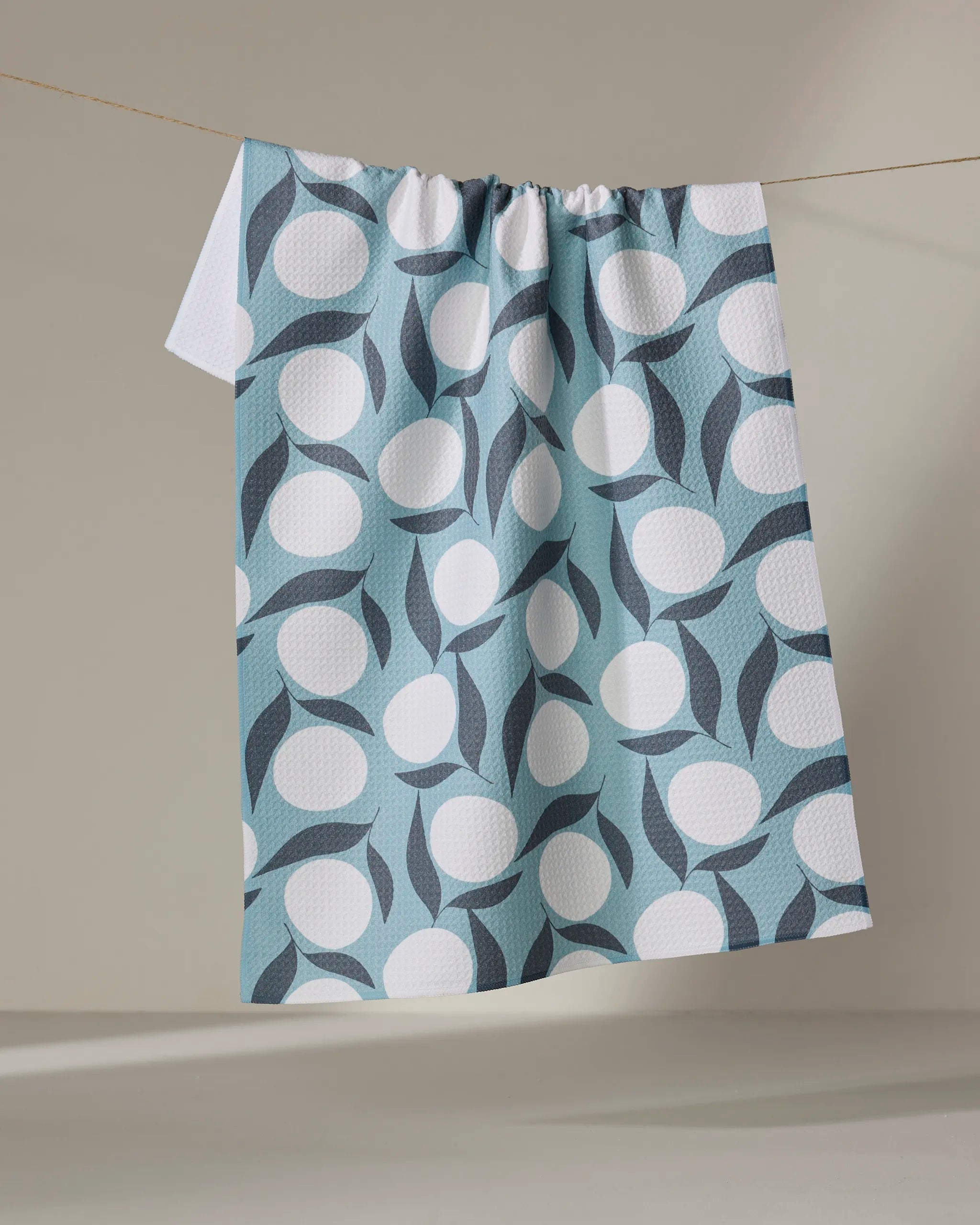 The Bouncing Blooms towel by Lizzie Clark, featuring a geometric design with large white circles and dark blue curves on light blue, hangs on a line against a neutral wall.