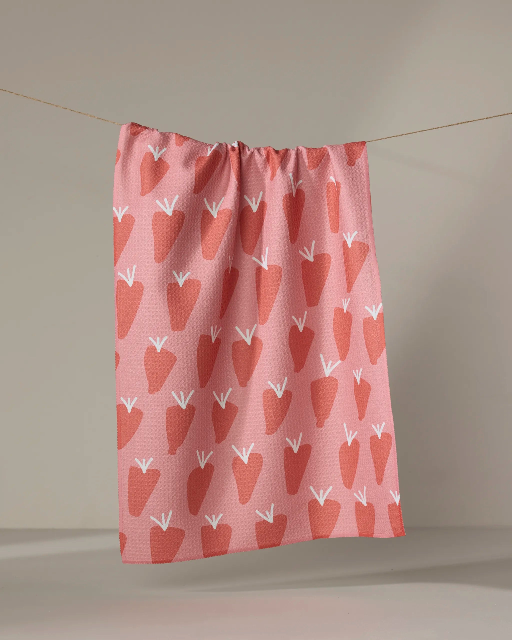 The Bountiful tea towel by Lizzie Clark, crafted from recycled material, displays a repeating pattern of stylized red strawberries with white stems on pink and hangs on a clothesline against a neutral background.
