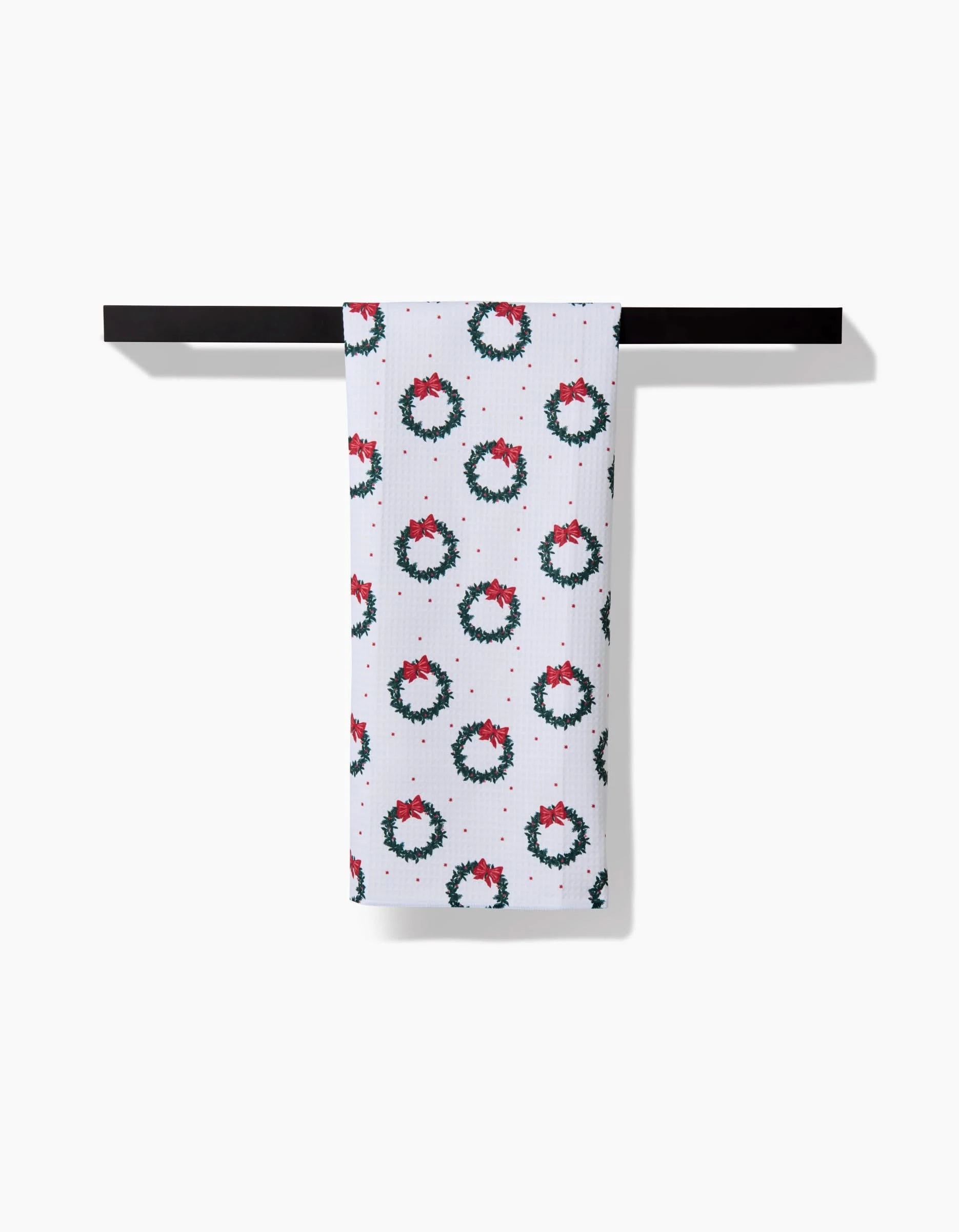 The Ash Cascade Bow Wreath white kitchen towel, made from recycled materials, displays green Christmas wreaths with red bows and hangs neatly on a black towel bar against a white background.
