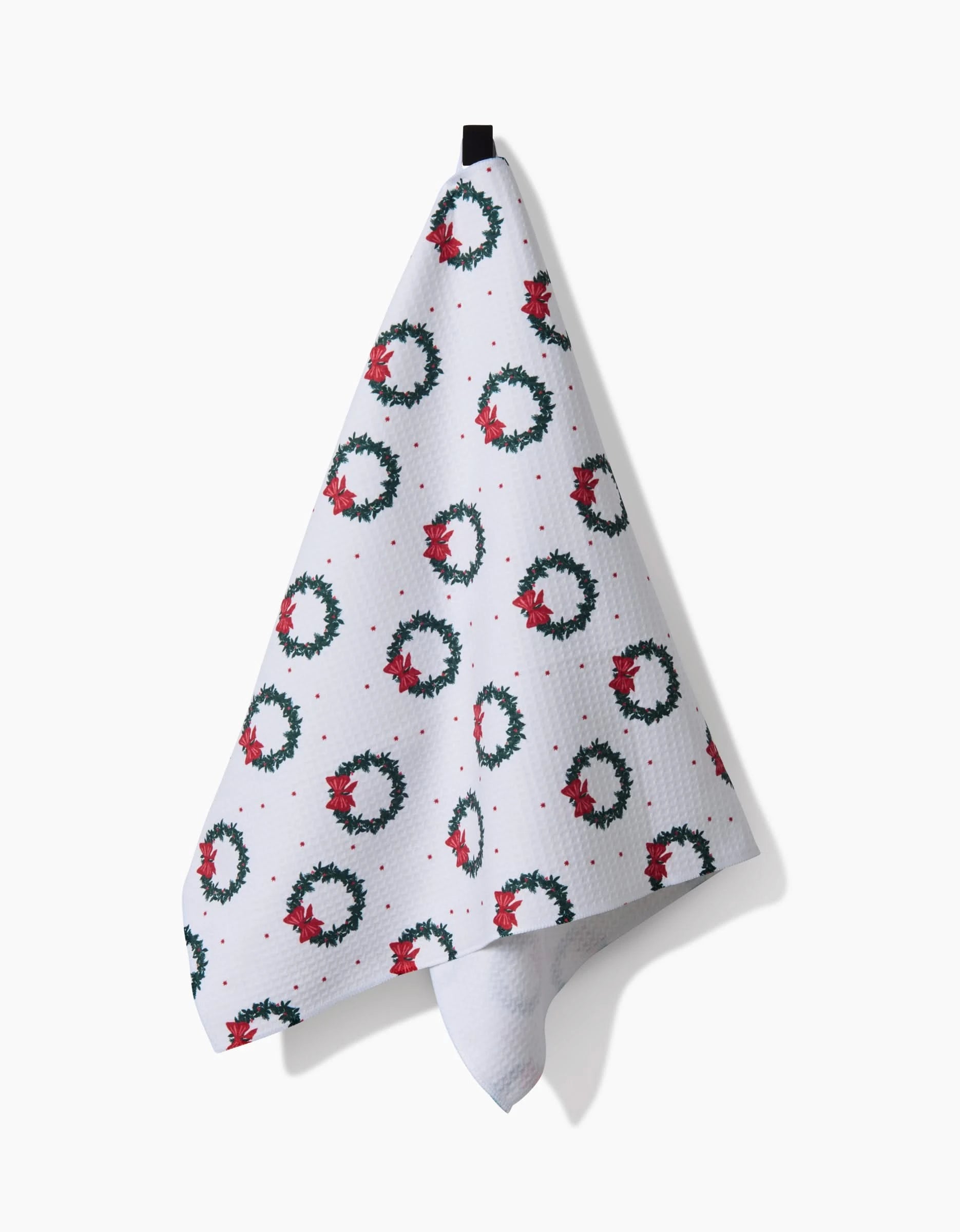 The Ash Cascade Bow Wreath kitchen towel features green holiday wreaths with red bows and berries on white fabric, hanging by a black loop against a plain white background.
