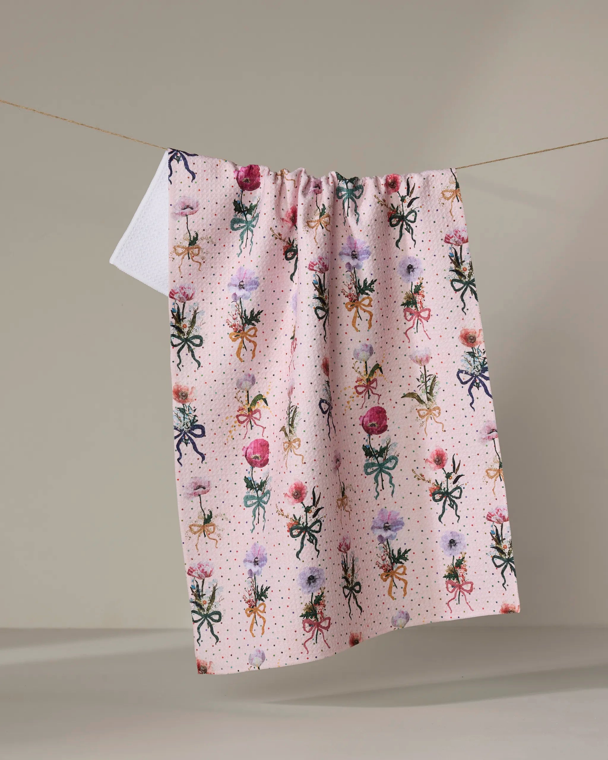 The Bows and Dots tea towel by Pascale Dilger, made from recycled materials and decorated with colorful octopuses and flowers, hangs on a clothesline against a neutral background.
