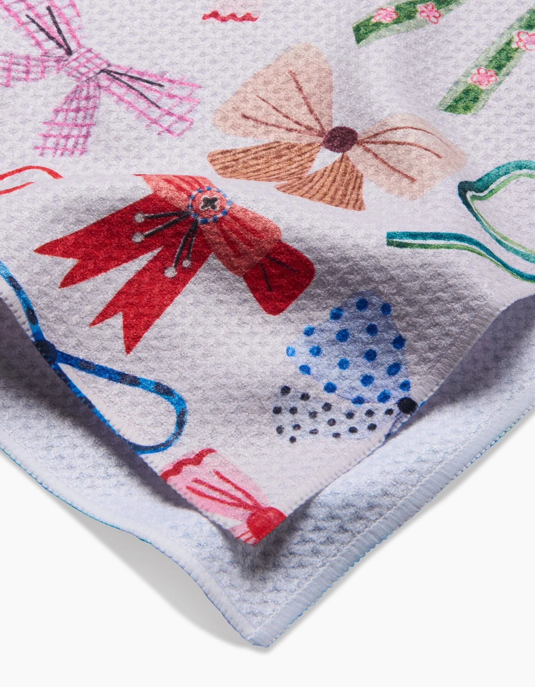 The “Bows” tea towel by Daria Solak is a textured white towel made from recycled materials, featuring colorful bow designs—red, blue polka dot, pink, brown, and green patterns—with one corner folded over.