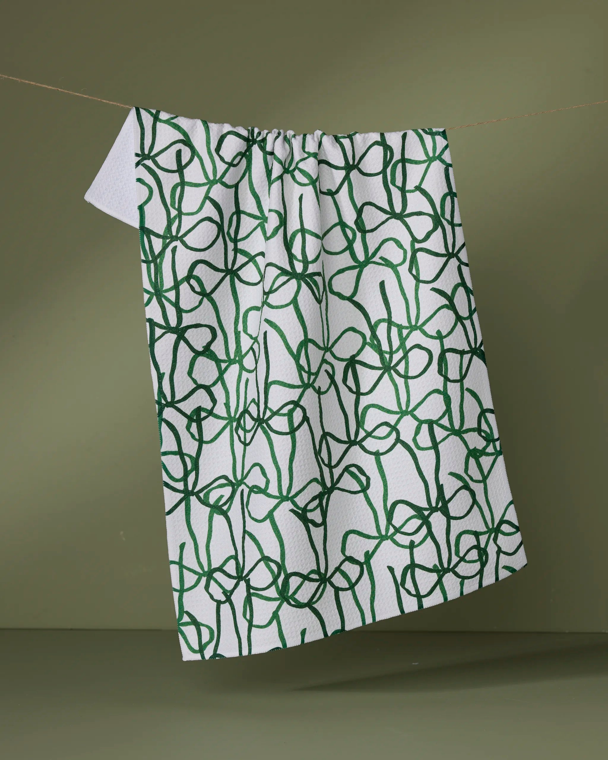 The Bowtiful kitchen towel by Geometry, featuring an abstract green line pattern on white, hangs on a clothesline against a plain olive green background.