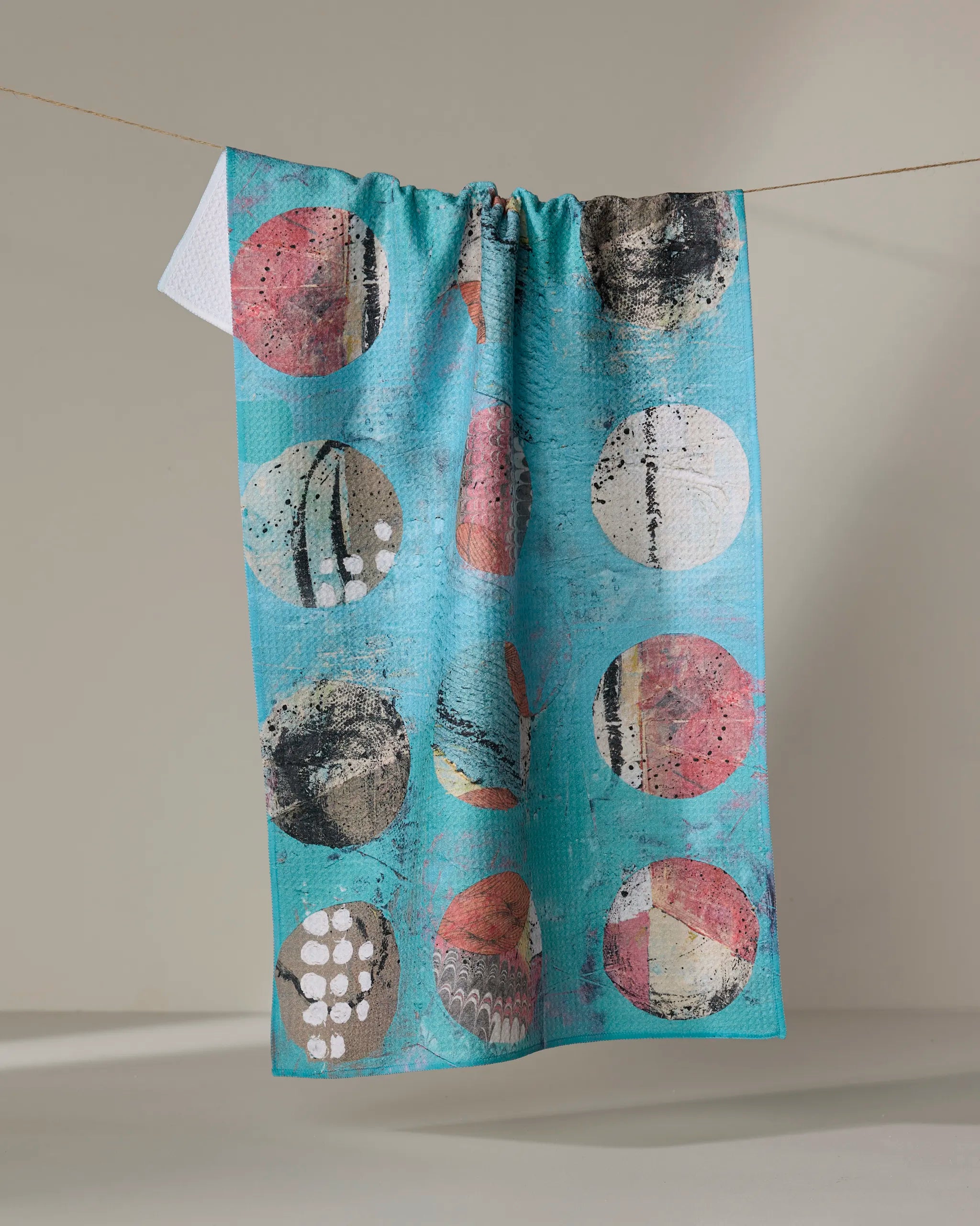 The "Boxed In" tea towel by Katherine Berkeley, featuring large, abstract multicolored circles and crafted from recycled materials, hangs on a thin clothesline against a neutral background.