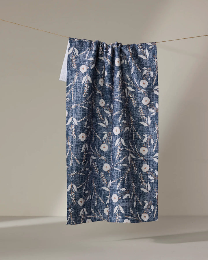 The Boyce Blue kitchen towel by Becki Owens features a blue and white floral pattern with subtle pink accents, hanging from a thin string against a plain, light-colored background.