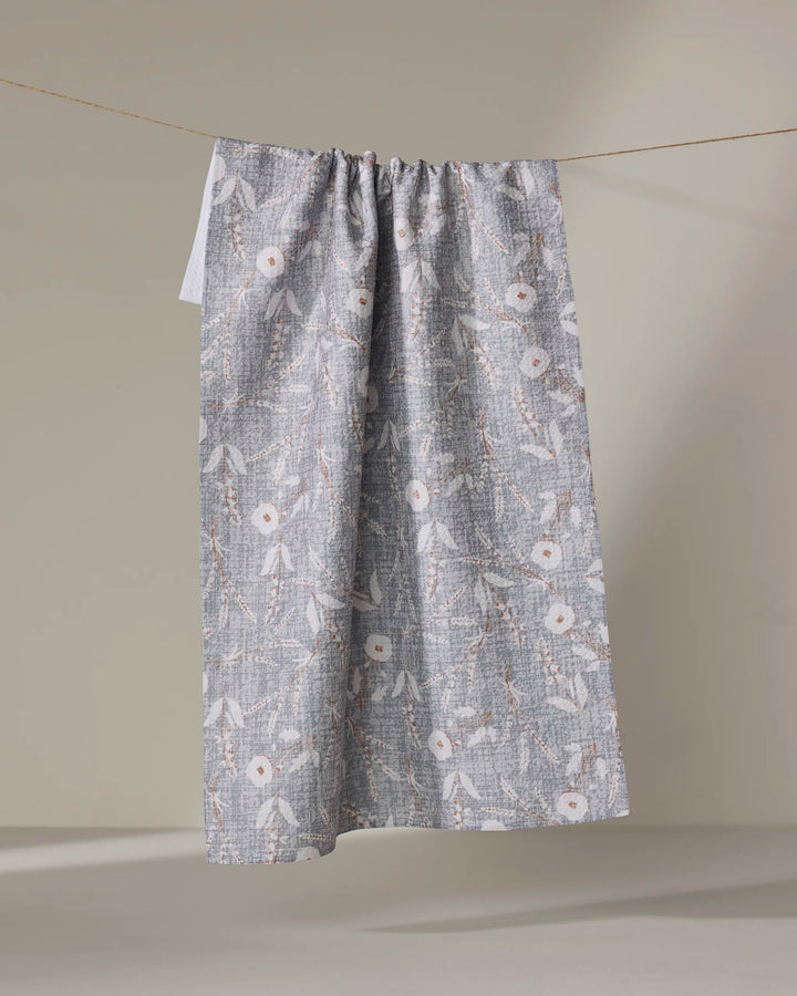 The Boyce Grey tea towel by Becki Owens, made from recycled materials and showcasing a white and beige floral pattern, hangs on a clothesline against a plain beige background.