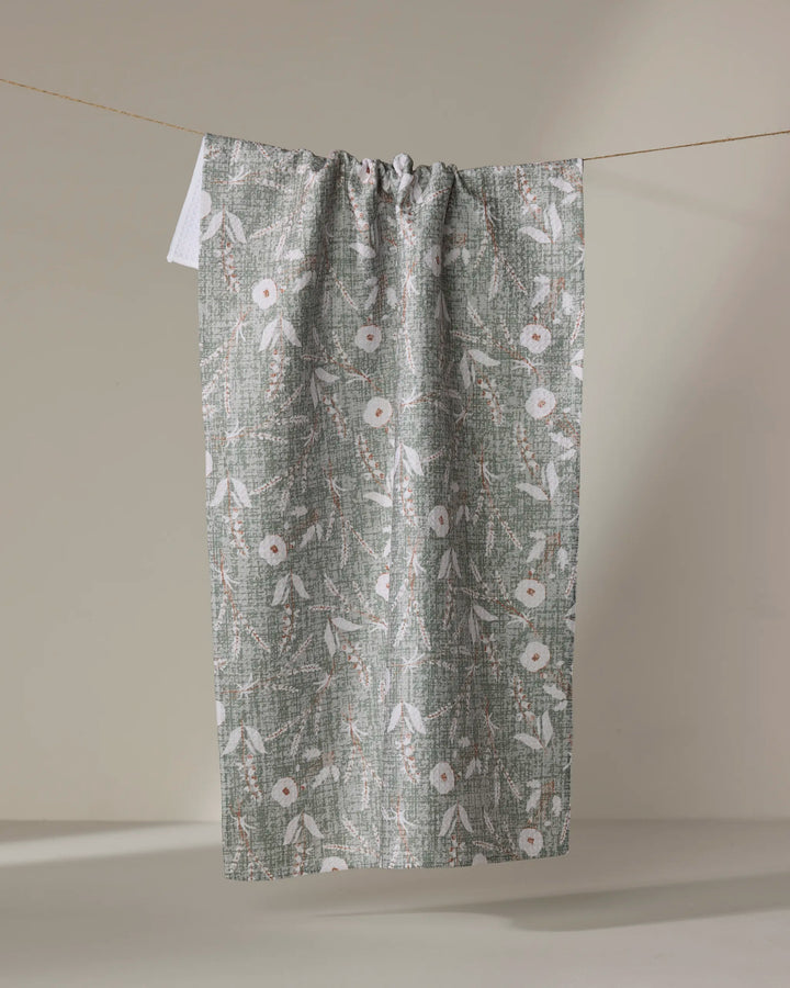 The Boyce Sage tea towel by Becki Owens, crafted from recycled materials, dries quickly and displays a white floral and leaf pattern. It is shown hanging on a string against a neutral beige background.