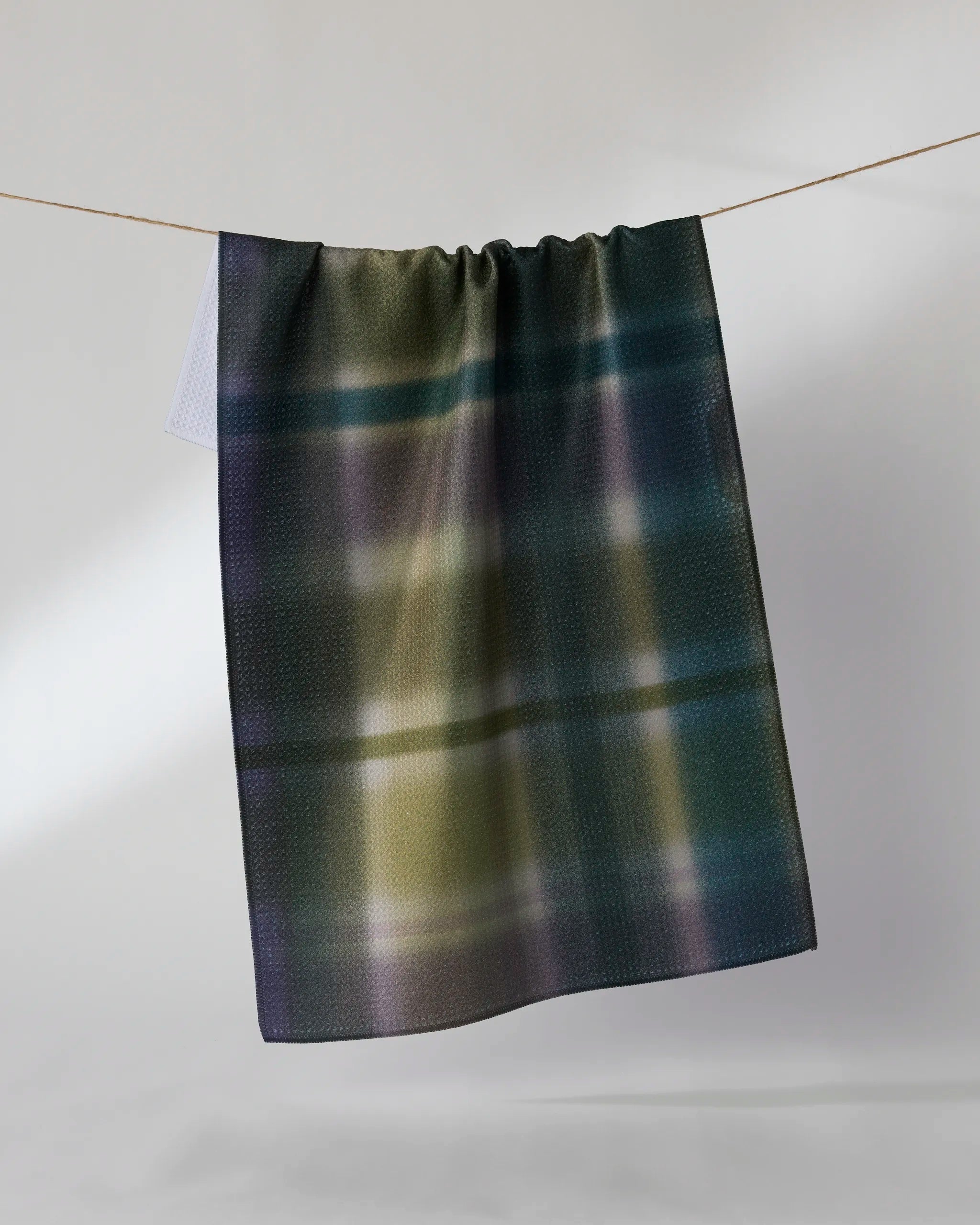 Bracken by Geometry is a soft, rectangular recycled kitchen towel featuring a dark gradient plaid in green, blue, and yellow tones, draped over a string with a light gray background.