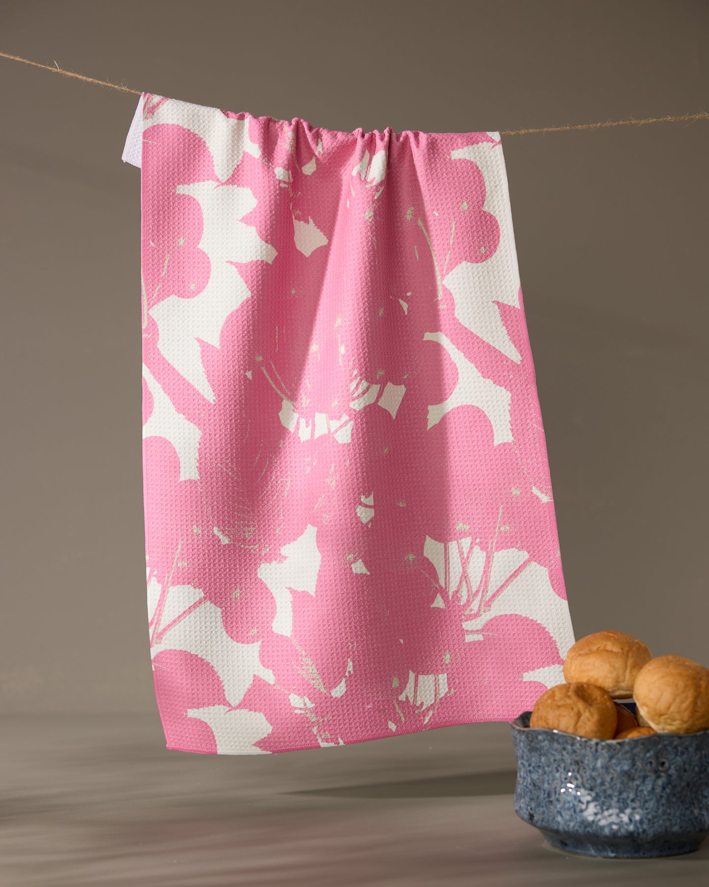 A Geometry Bramble Pink tea towel with pink floral patterns hangs on a clothesline. In front, a blue ceramic bowl filled with walnuts stands out against a softly lit, neutral background.
