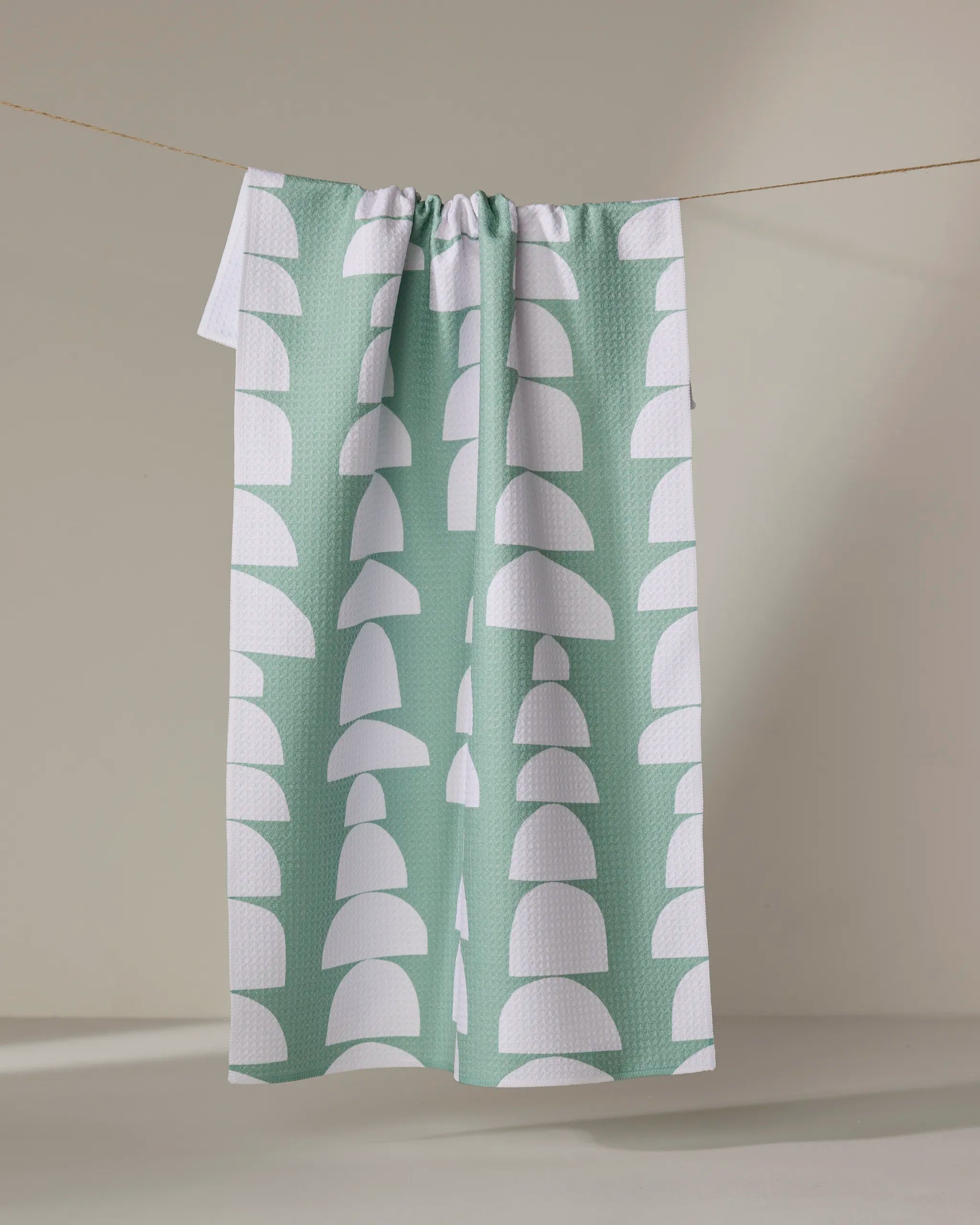 The "Breakfast at Granny's" dish towel by Claire Iglesias, in sage green with white semi-circle and arch patterns, hangs on a clothesline against a neutral background.
