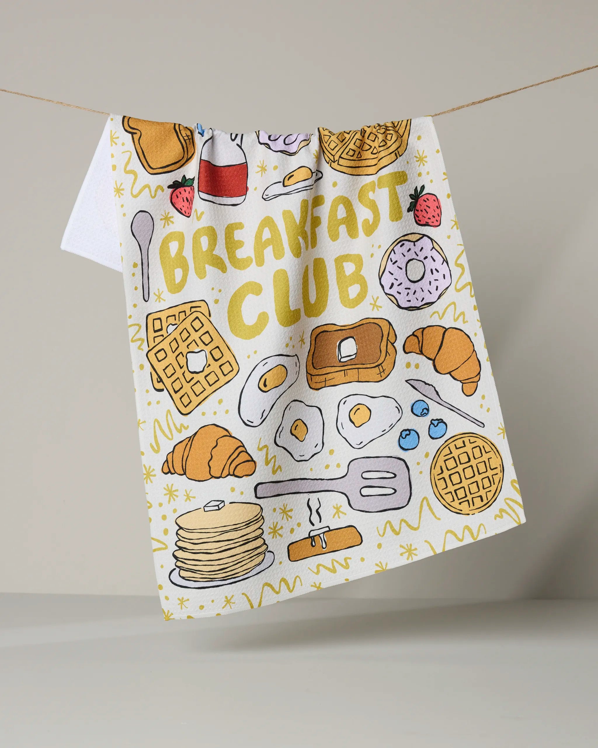 The Breakfast Club tea towel by Rosa Kichline features bold "BREAKFAST CLUB" lettering and playful breakfast illustrations, adding color and charm to your kitchen while providing absorbent practicality.