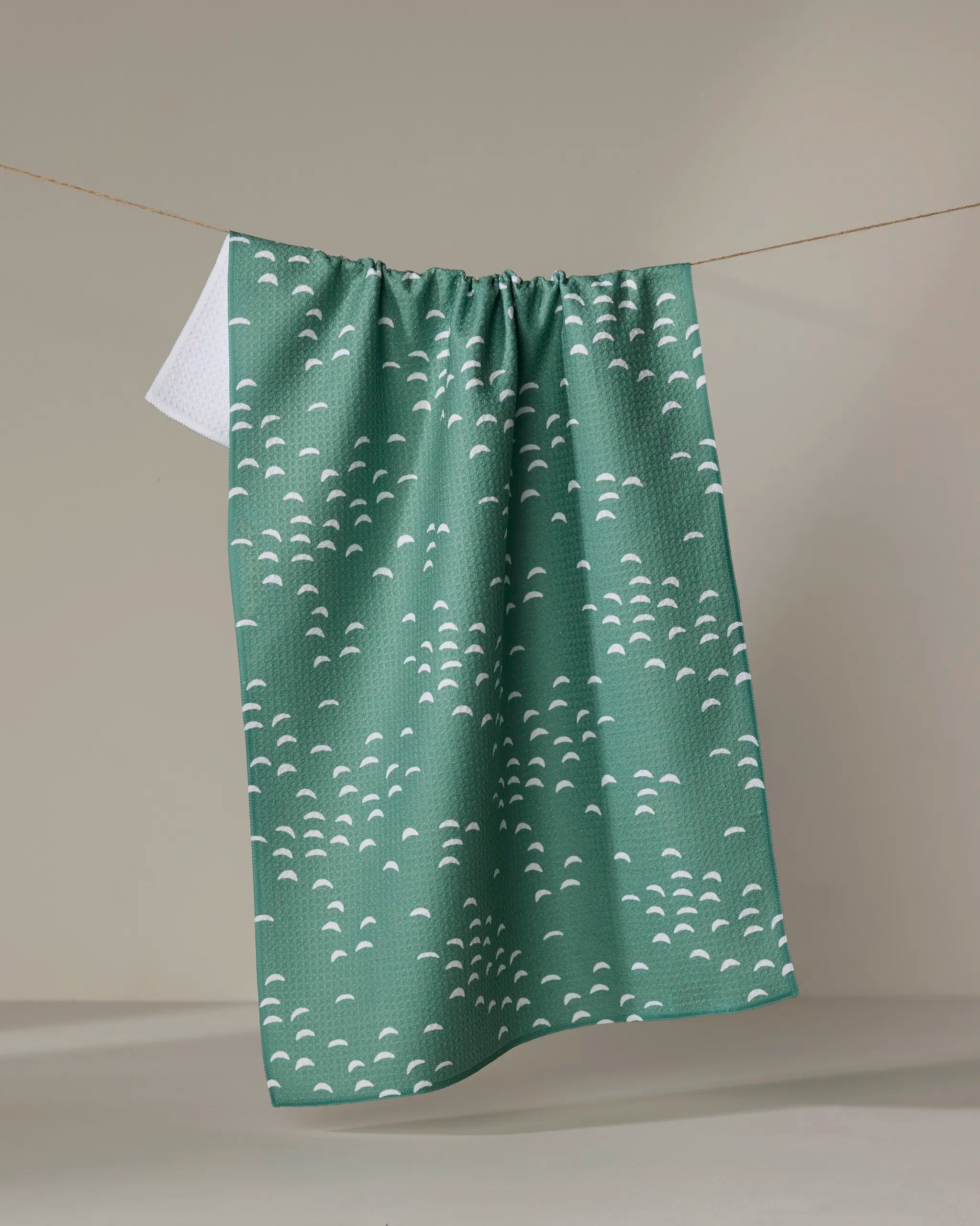 The Breezeway kitchen towel by Jessica Swift features a green color with small white semi-circles, crafted from recycled materials, hanging on a clothesline against a beige background.