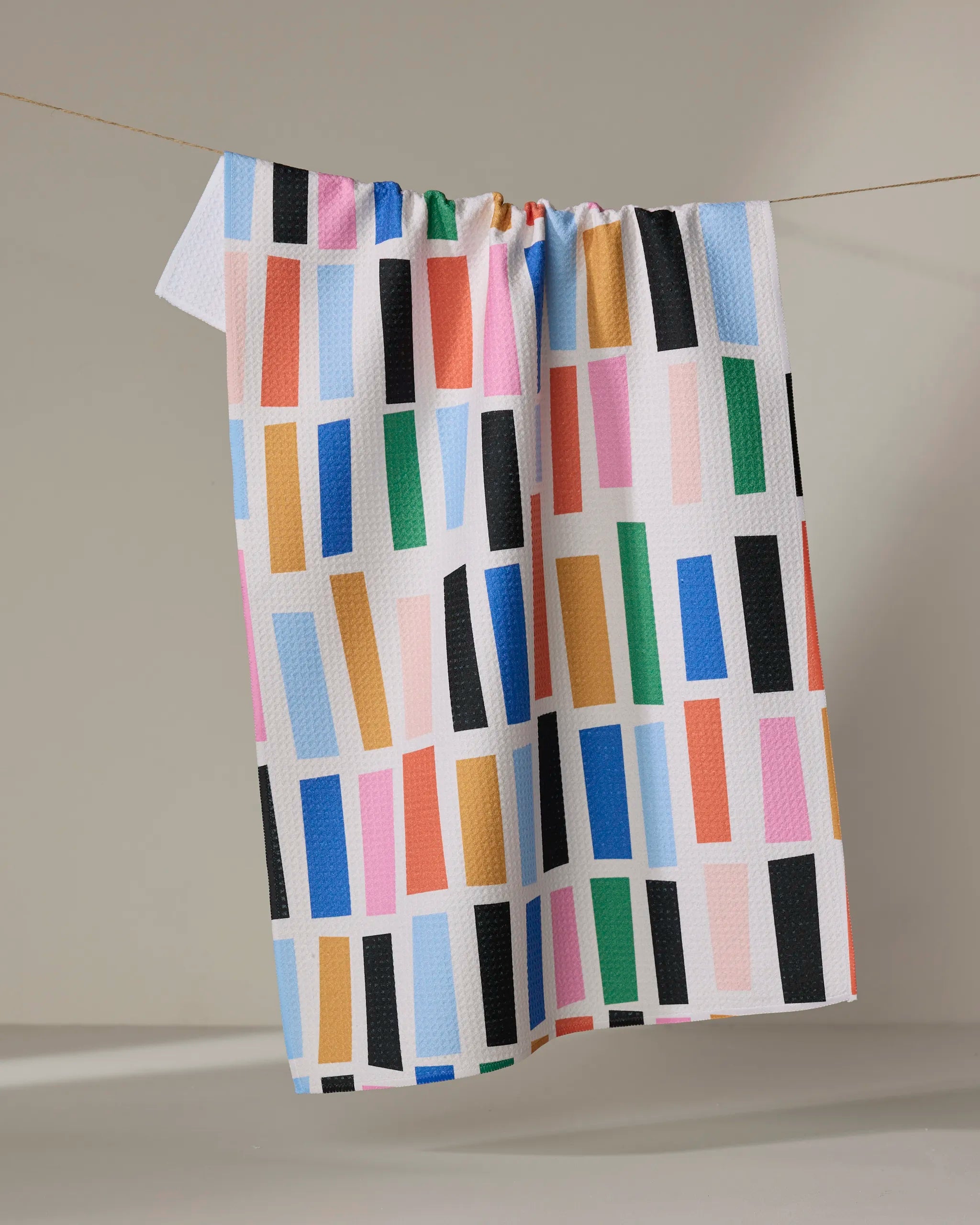 The Bricks tea towel by Jennifer Bouron features a colorful geometric pattern of vertical rectangles in blue, pink, orange, green, and black. Made from recycled materials, it dries quickly and adds sustainable style to any kitchen.
