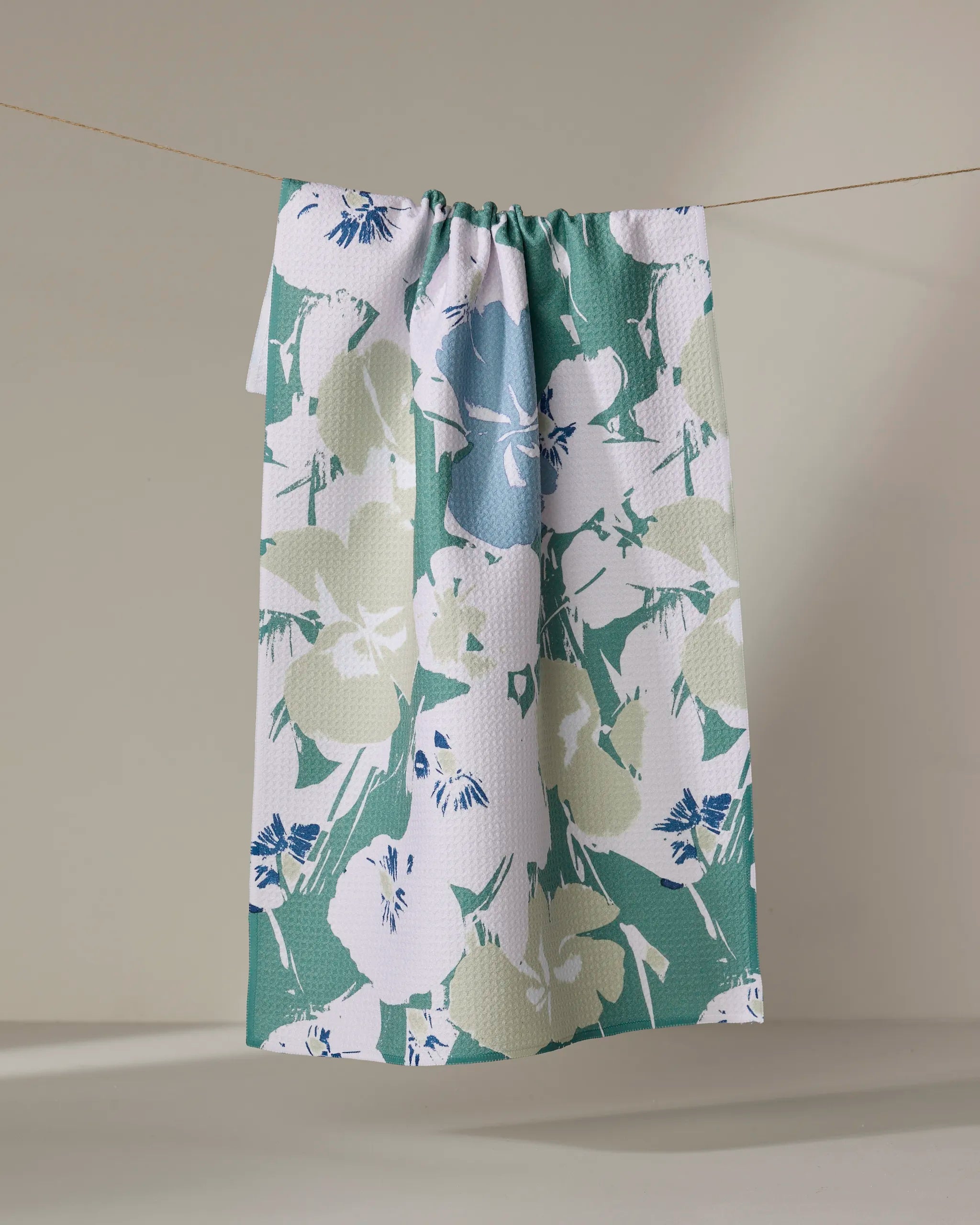 The Geometry Brie towel, featuring a green, white, beige, and blue floral pattern and quick-drying textured fabric, hangs draped over a clothesline against a neutral background.