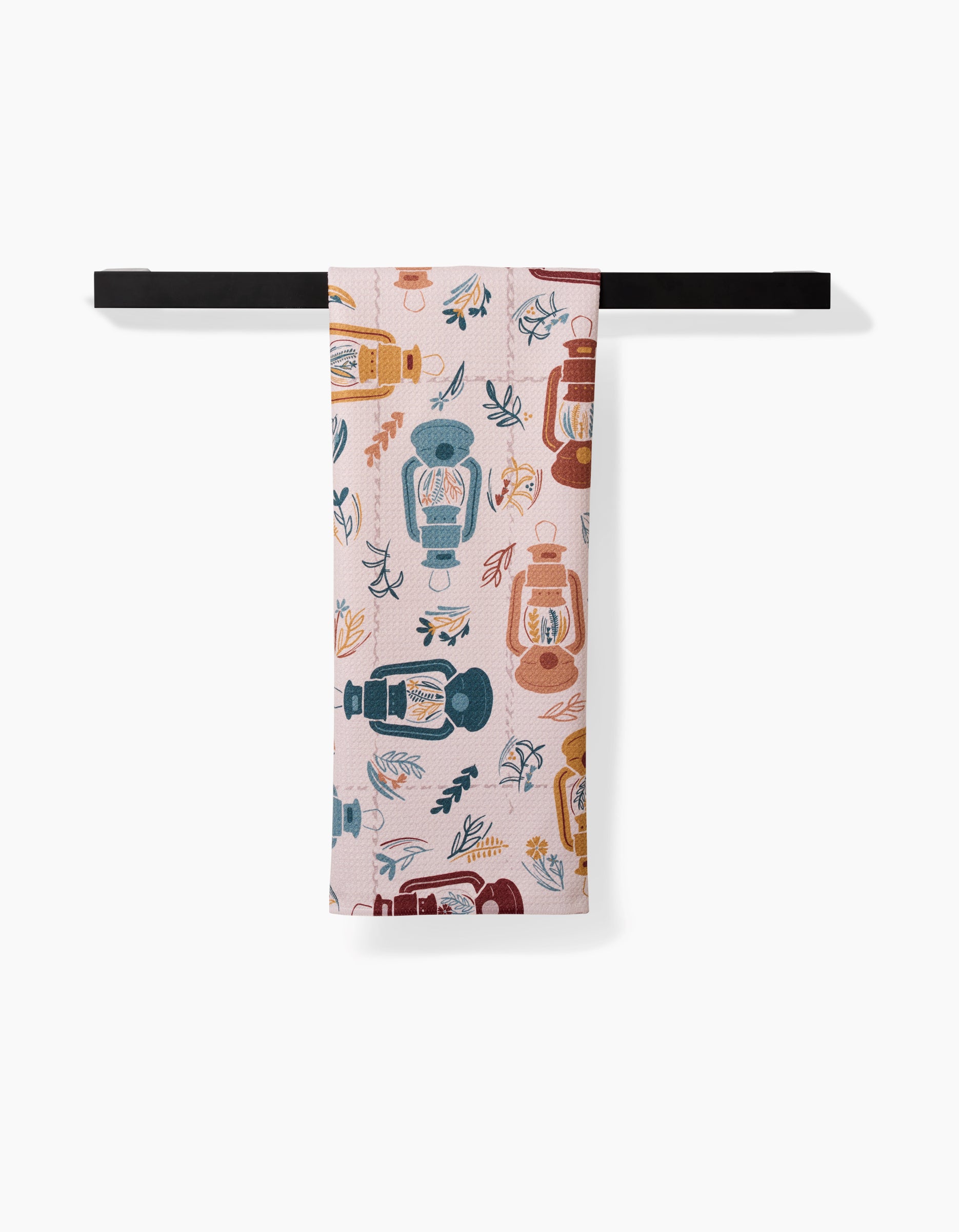 The Twig + Spark Bright Spot tea towel, featuring a geometric lantern and leaf pattern in blue, orange, and red, hangs neatly folded over a horizontal black bar against a white background.