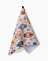 The Bright Spot tea towel by Twig + Spark features lantern, leaf, and branch illustrations in blue, brown, and mustard on pale pink—crafted from recycled materials for an eco-friendly kitchen accent.