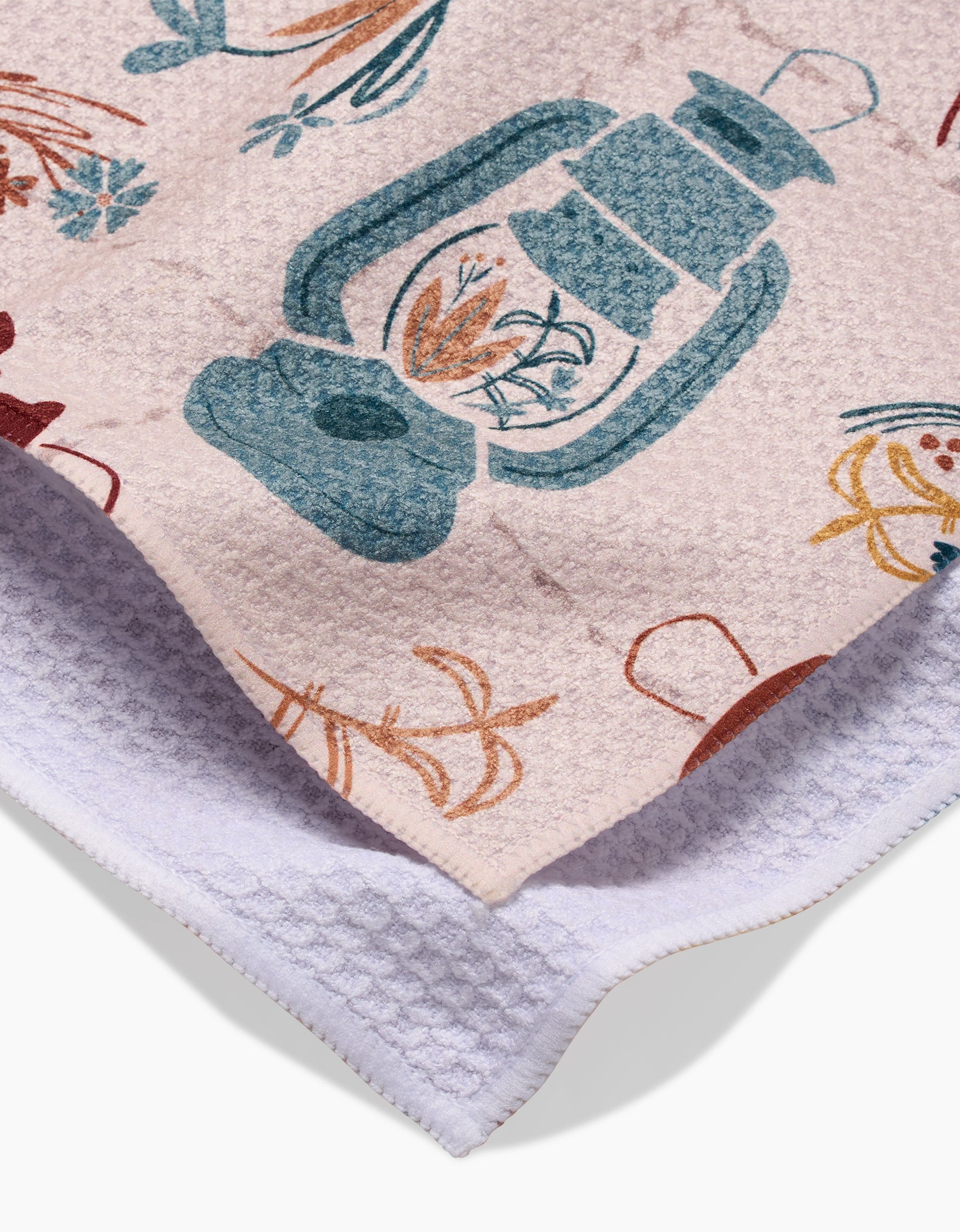 A close-up of the Bright Spot tea towel by Twig + Spark, made from recycled materials, shows its white textured fabric with blue lanterns and orange-brown florals printed on one side; its folded to display both the print and plain sides.