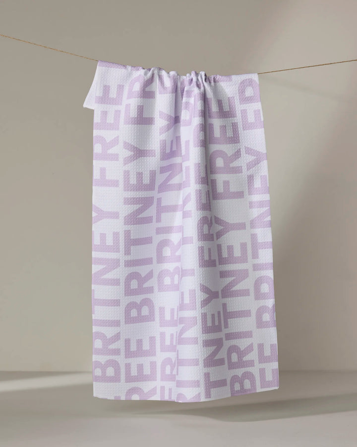 The Buy Guide’s “Britney” tea towel, made from recycled materials, features FREE BRITNEY in bold light purple lettering and hangs on a clothesline against a neutral background.