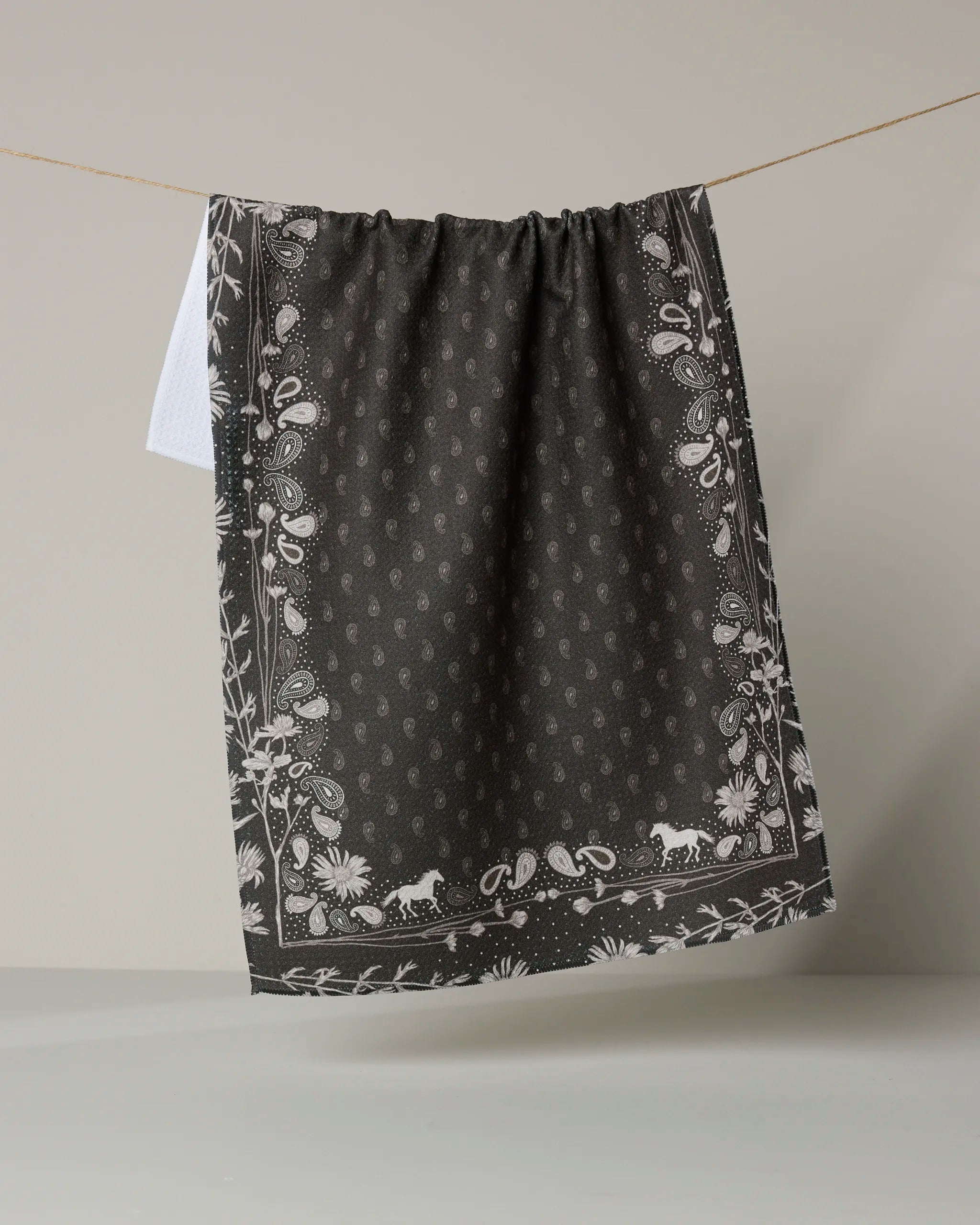 The Geometry Bronco Espresso tea towel features a white paisley and floral border on black, hanging against a plain light background—an absorbent dish towel ideal for any kitchen.