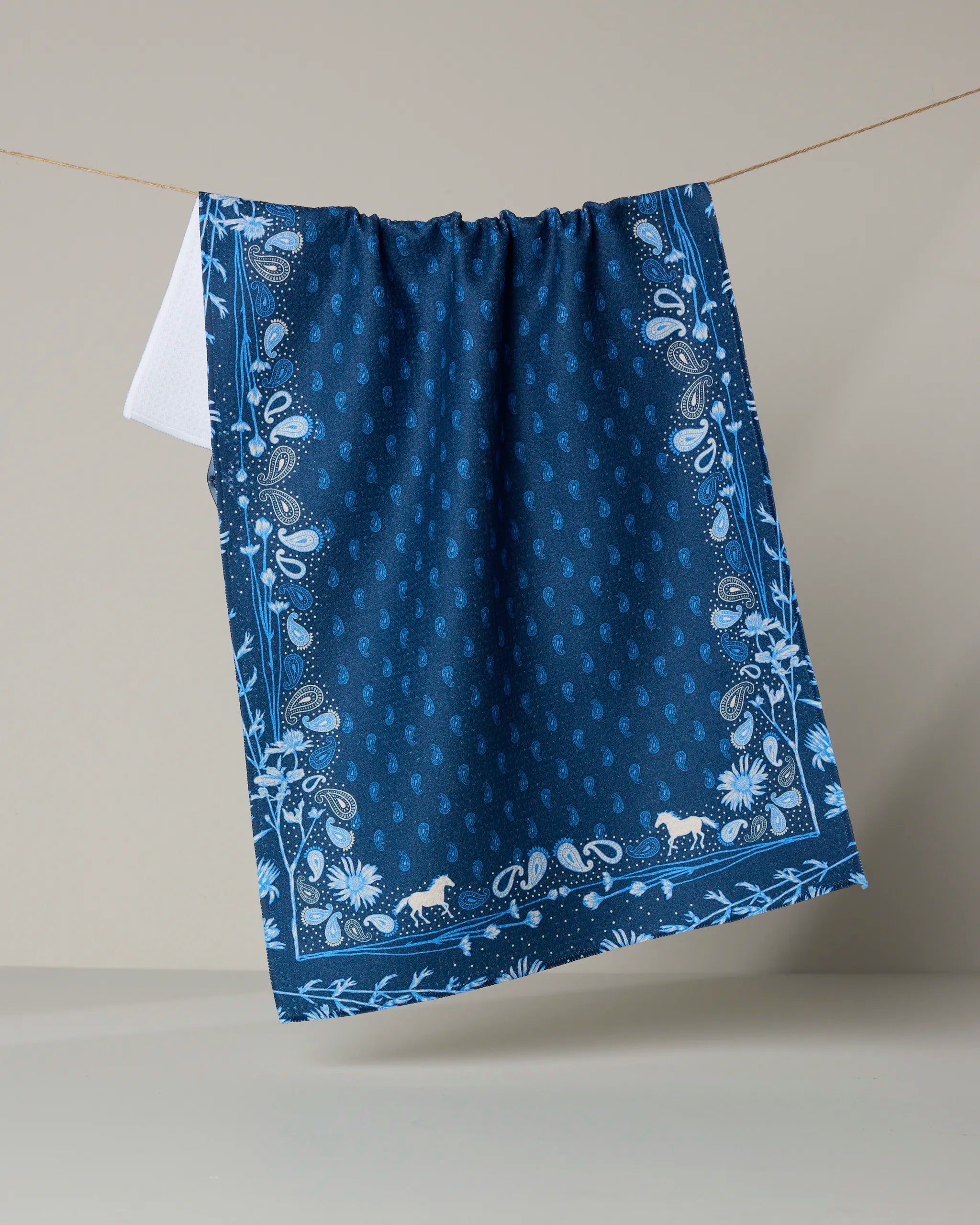 The Geometry Bronco Indigo dish towel features a dark blue, absorbent fabric with a white paisley and floral border plus small animal silhouettes, shown hanging on a string against a plain light gray background.