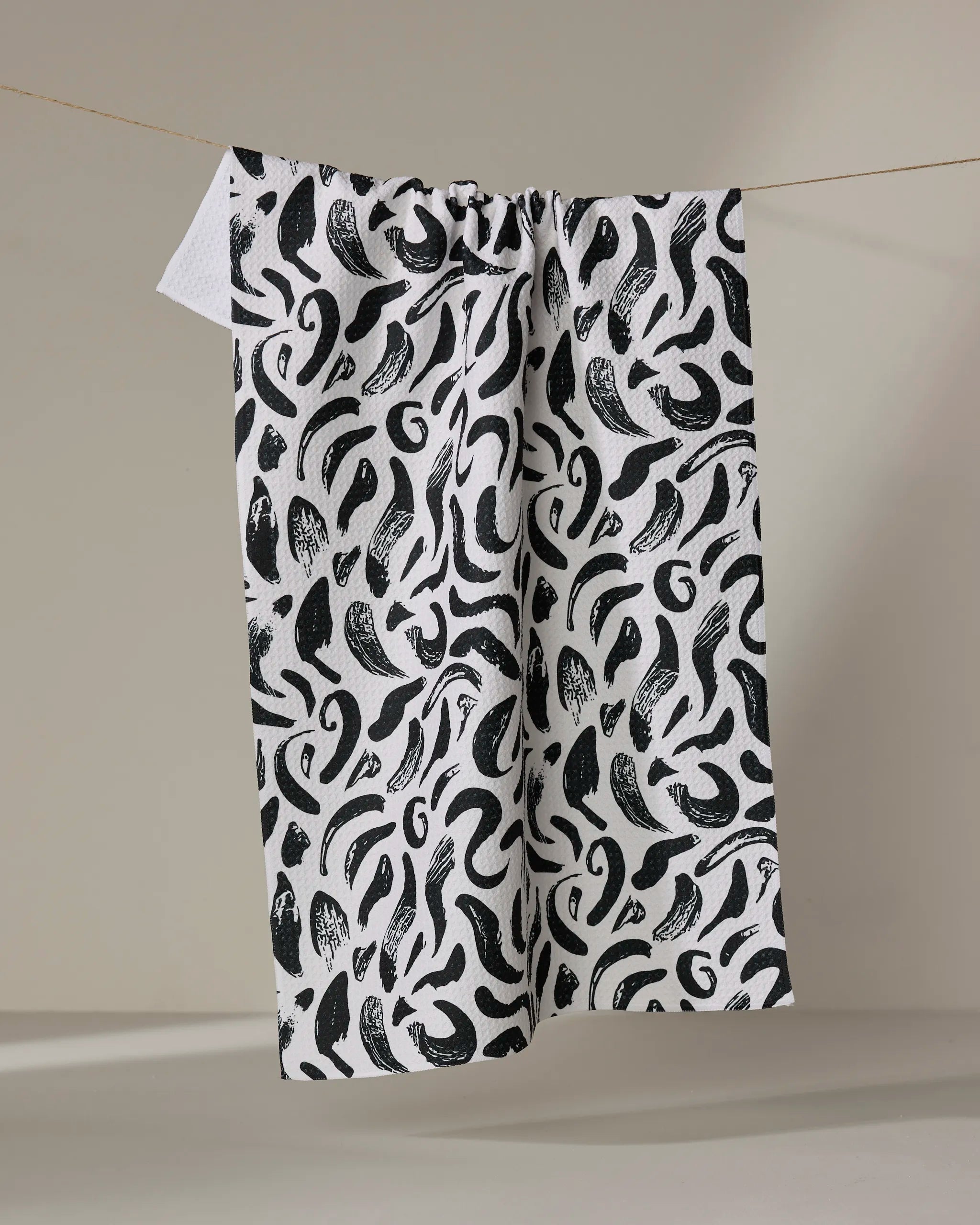 The Brush Strokes Swirls tea towel by Sarah Daugherty, crafted from recycled materials, features a bold black abstract pattern and hangs on a thin clothesline against a neutral background.