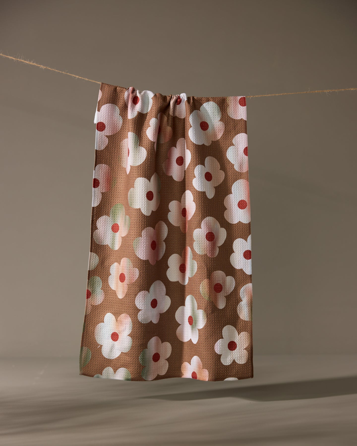 The Geometry Brushed Bouquet Brown kitchen towel, featuring a pattern of large white and pink flowers, hangs against a plain beige background.