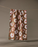 The Geometry Brushed Bouquet Brown kitchen towel, featuring a pattern of large white and pink flowers, hangs against a plain beige background.
