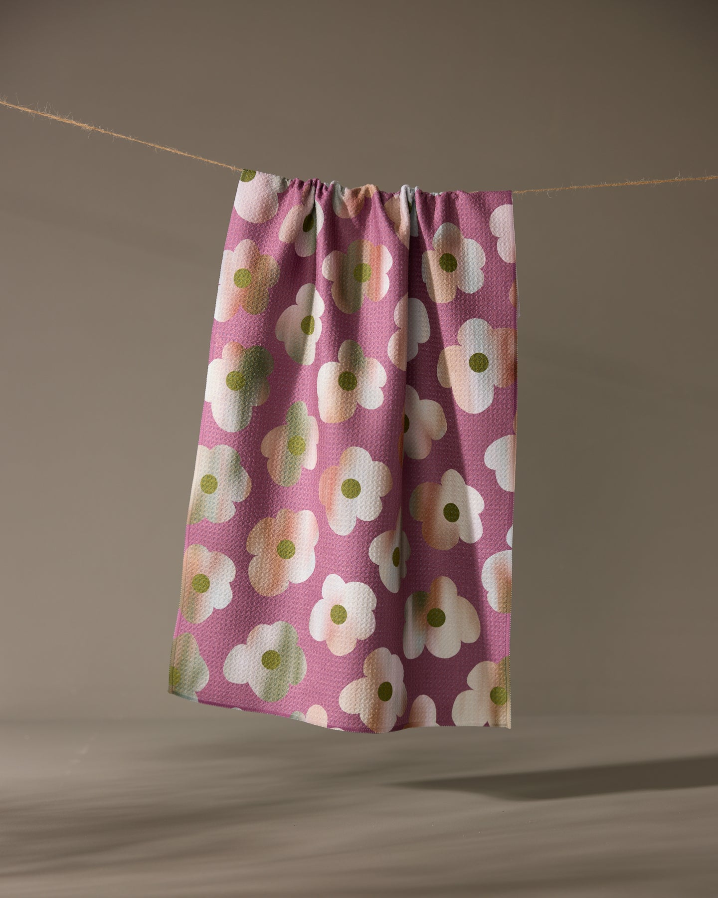 The Brushed Bouquet tea towel by Geometry, featuring bold white and green florals on pink and made from recycled materials, hangs from a clothesline against a neutral background.