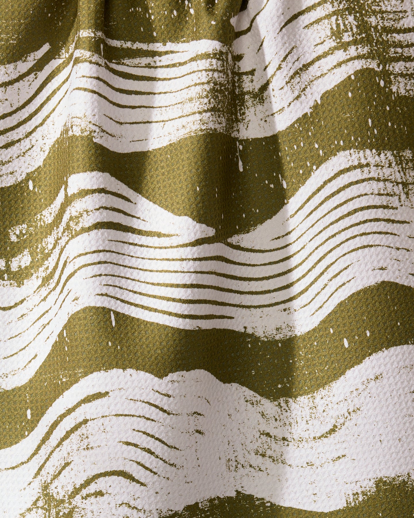 Close-up of Geometrys Brushline Green ultra-absorbent towel, showcasing an abstract pattern of wavy, irregular white lines of varying thickness on an olive green background. The fabric has a slightly wrinkled appearance.