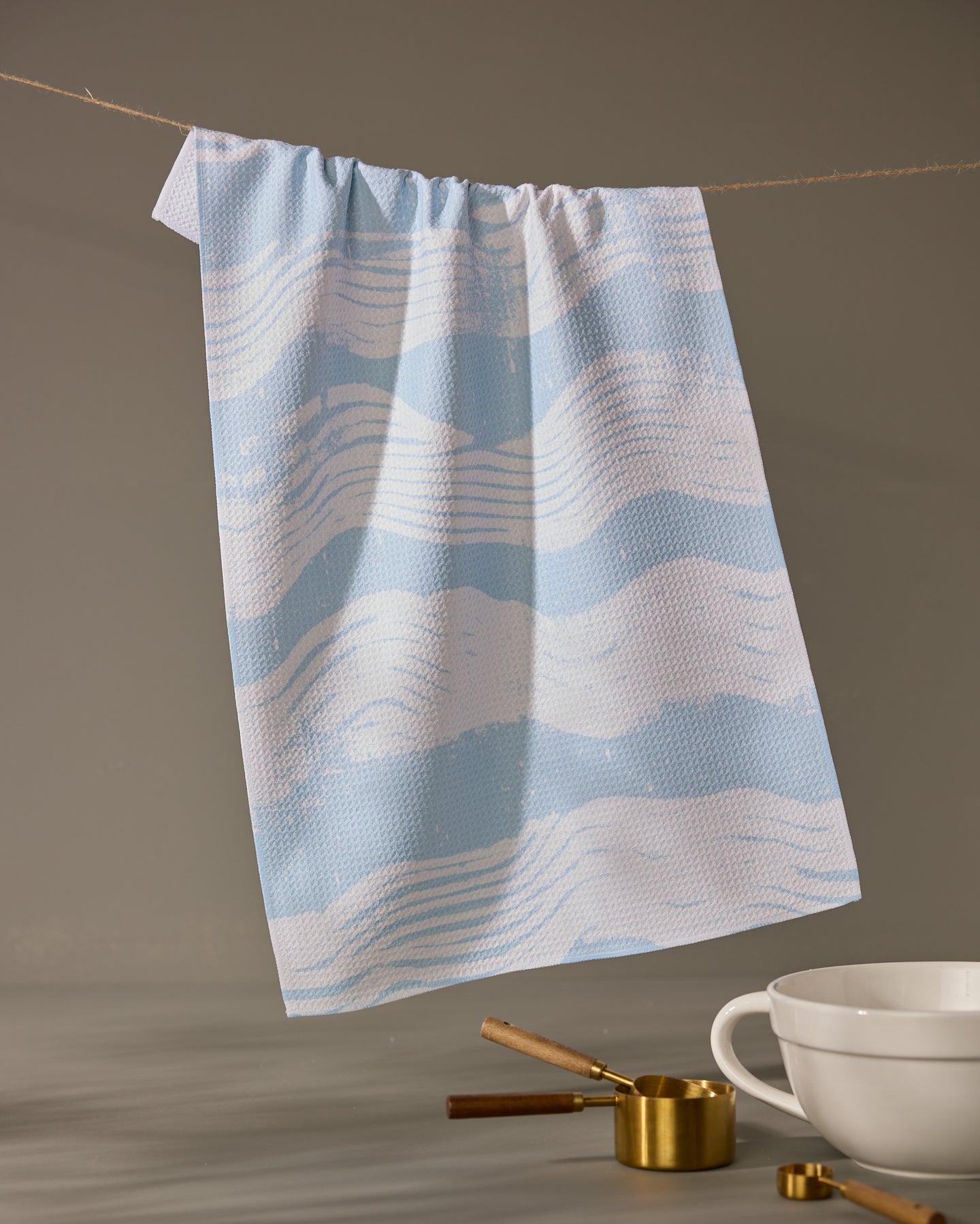 The Geometry Brushline Lightblue tea towel hangs on a string against a neutral background, with a white bowl, gold measuring cups, and a wooden spoon arranged below.