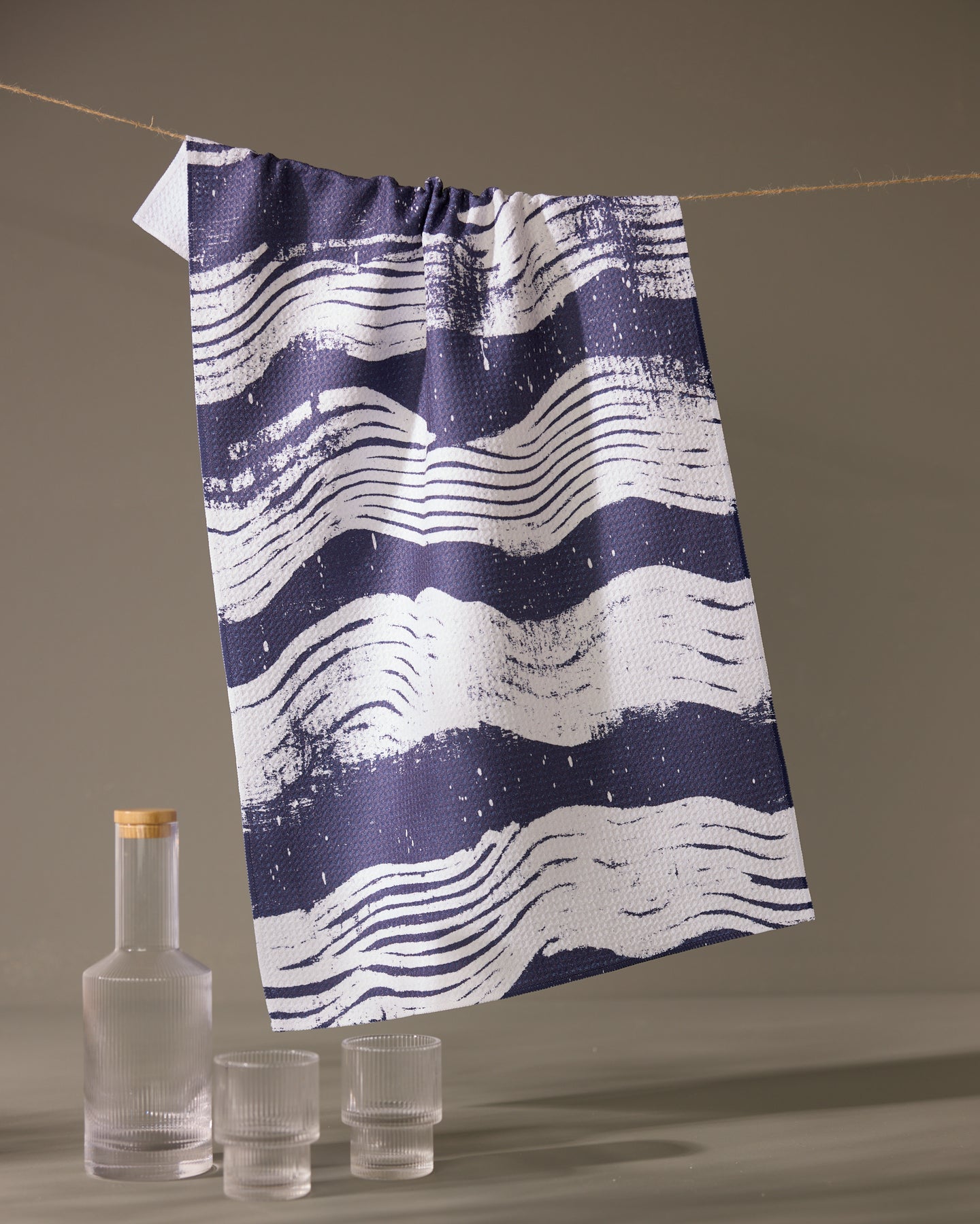 The Geometry Brushline Navy quick-drying tea towel, featuring dark blue and white wavy stripes, hangs on a string above a glass bottle with a wooden cap and two matching textured glasses against a plain backdrop.