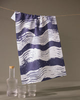 The Geometry Brushline Navy quick-drying tea towel, featuring dark blue and white wavy stripes, hangs on a string above a glass bottle with a wooden cap and two matching textured glasses against a plain backdrop.