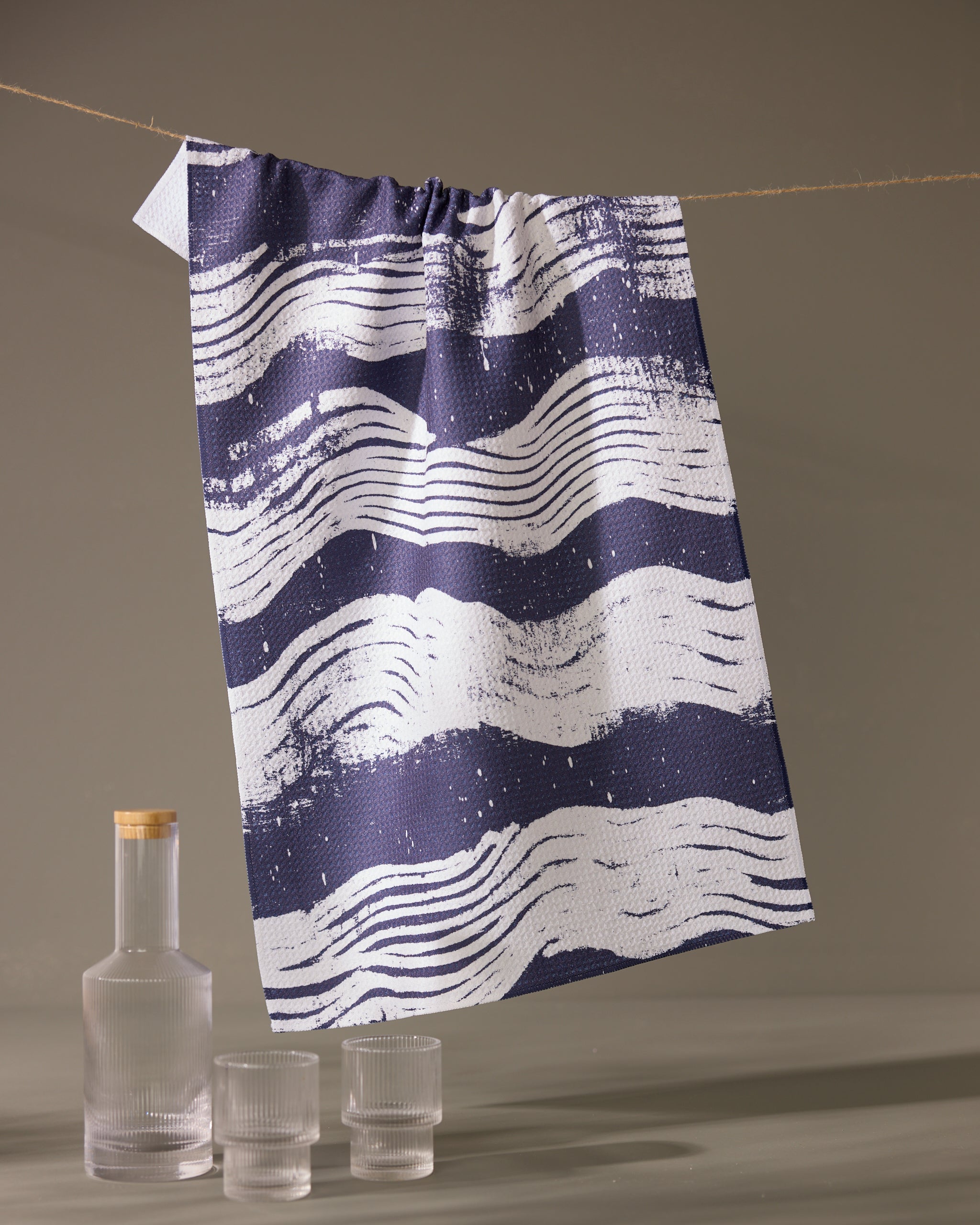 The Geometry Brushline Navy quick-drying tea towel, featuring dark blue and white wavy stripes, hangs on a string above a glass bottle with a wooden cap and two matching textured glasses against a plain backdrop.