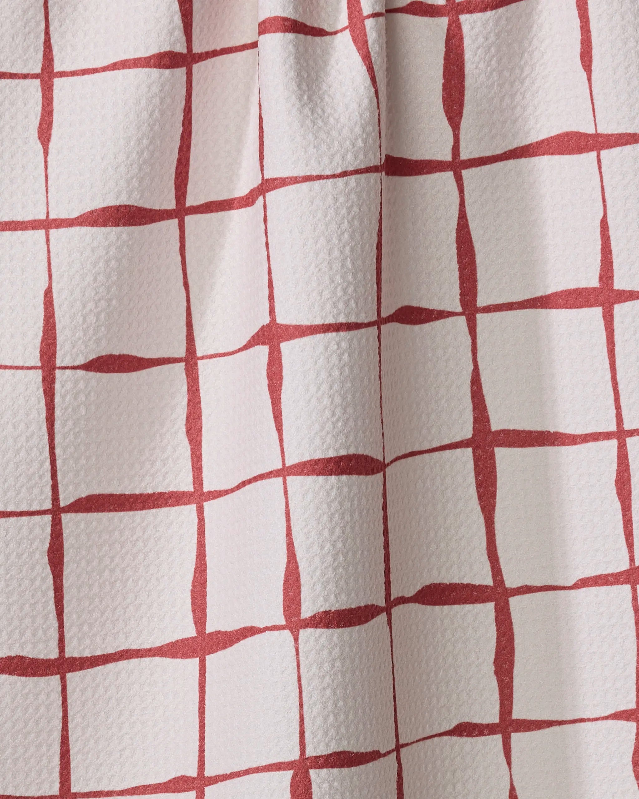 A close-up of the "Brushstroke Checks" white kitchen tea towel by Linda Fox, made from recycled materials, featuring a textured surface with a red irregular grid pattern, and visible fabric folds and shadows.