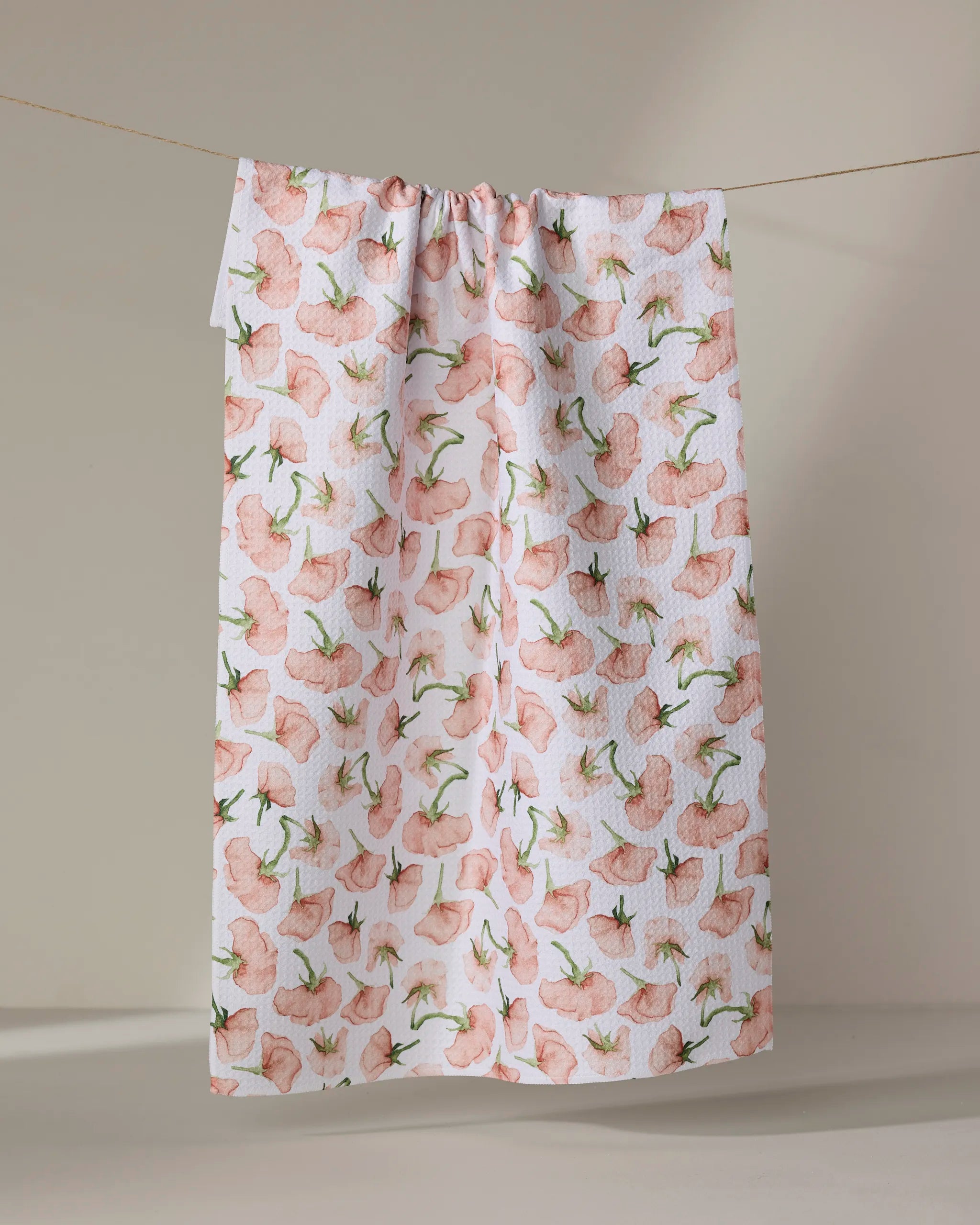 The "Budding Romance" tea towel by Aubrey Fairchild, featuring light pink flowers and green stems, hangs on a clothesline against a neutral background. Eco-friendly design.