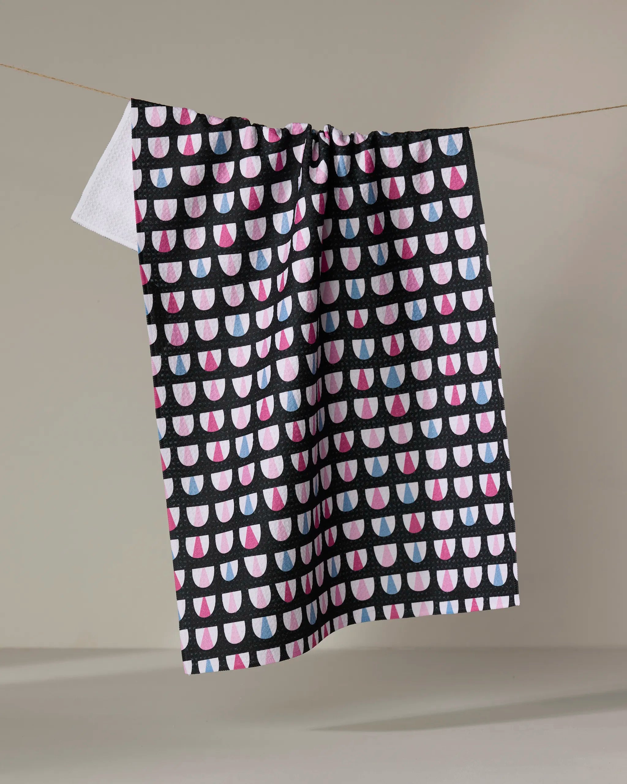 The Bug Boogie recycled kitchen towel by Anette Heiberg, with a geometric pattern of pink, white, and blue semi-circles on black, hangs on a line against a plain beige backdrop.