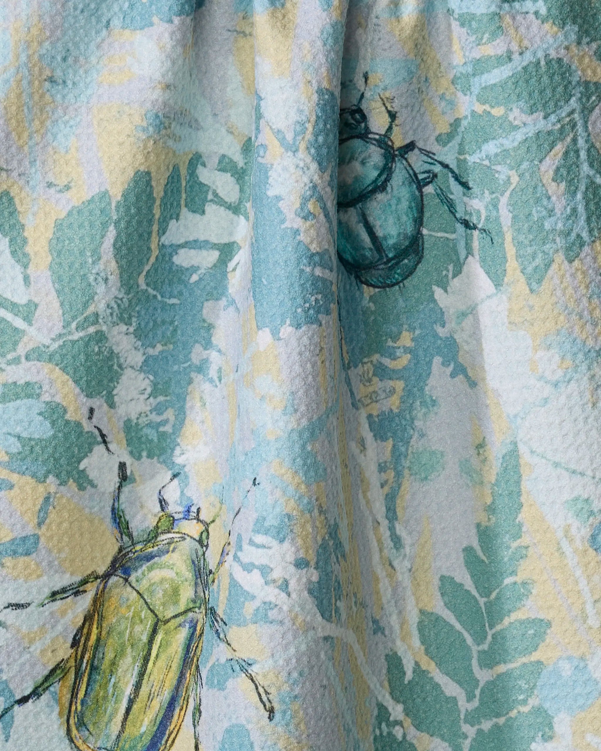 The "Bug Leaves" tea towel by Taryn Brennan features artistic green and yellow leaves with illustrated green and blue beetles, and is crafted from recycled materials.