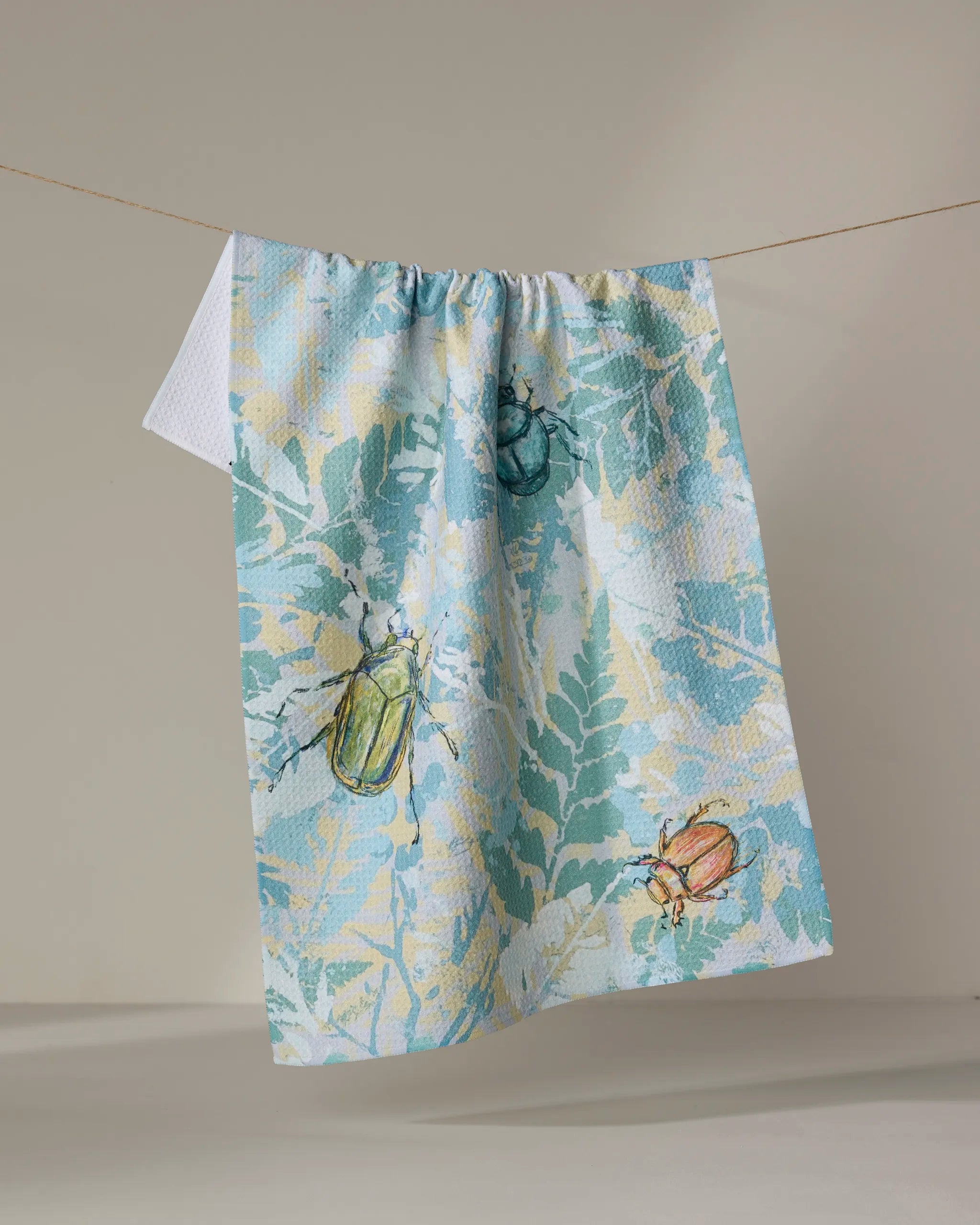 The Bug Leaves tea towel by Taryn Brennan, made from recycled materials, displays blue and yellow fern leaves with vibrant beetle designs and hangs on a clothesline against a plain beige backdrop.