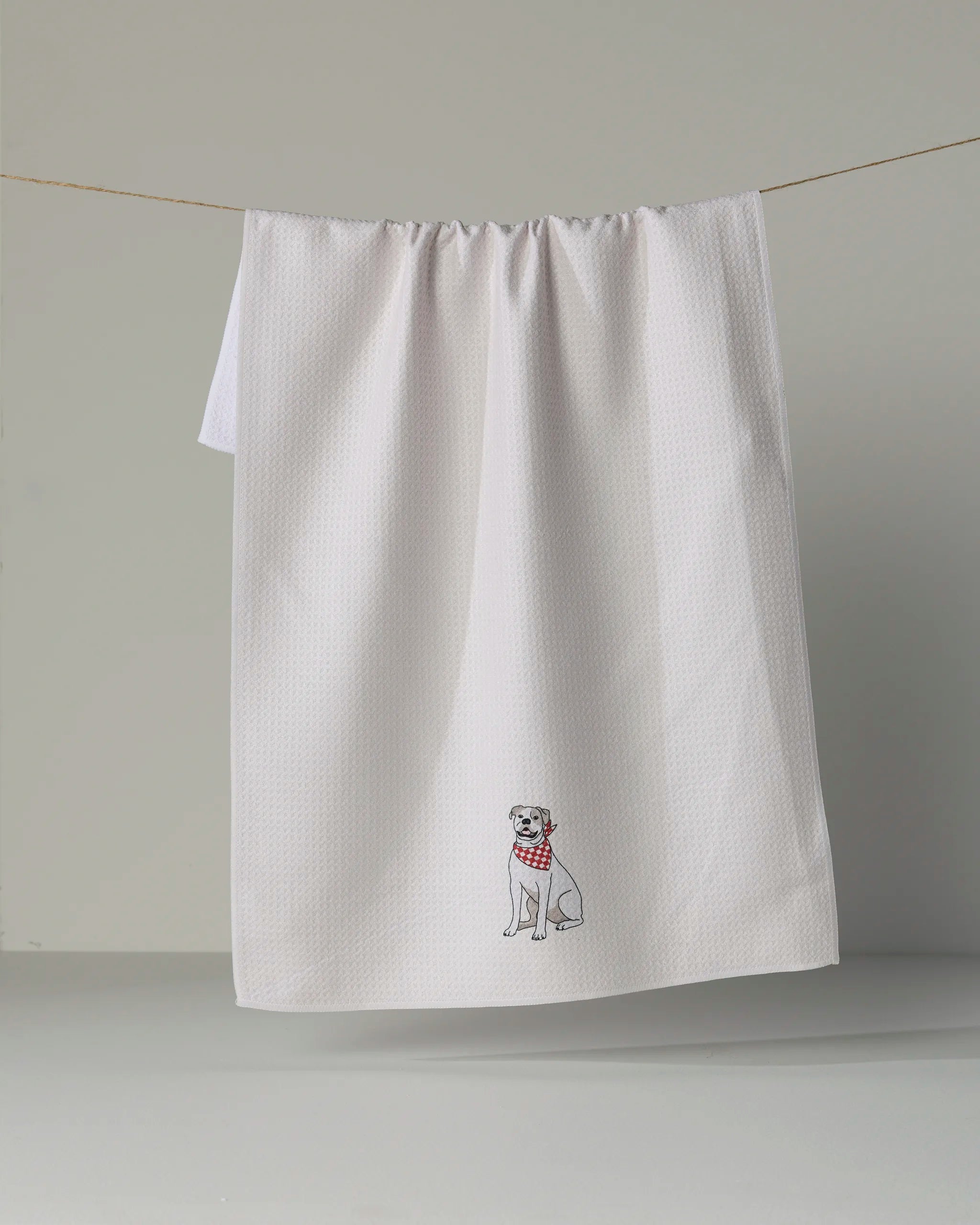 A white, eco-friendly “Bulldog Bandana” kitchen towel by Geometry hangs on a line, showcasing a small embroidered dog wearing a red checkered bandana near the bottom corner.