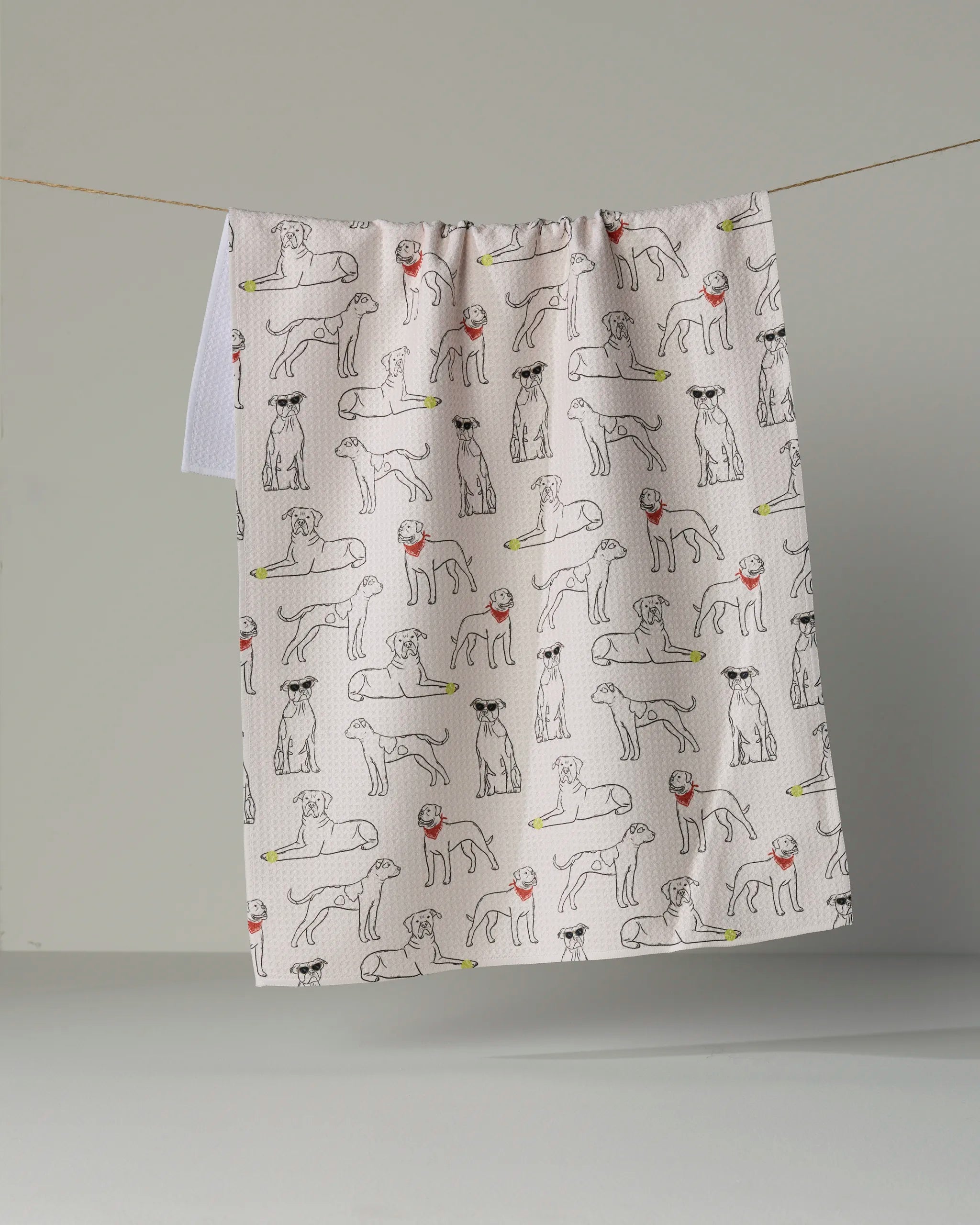 The “Bulldog Buddies” dish towel by Geometry features a white, absorbent fabric adorned with black line illustrations of different dog breeds sporting colorful sunglasses, displayed hanging on a clothesline against a neutral gray backdrop.