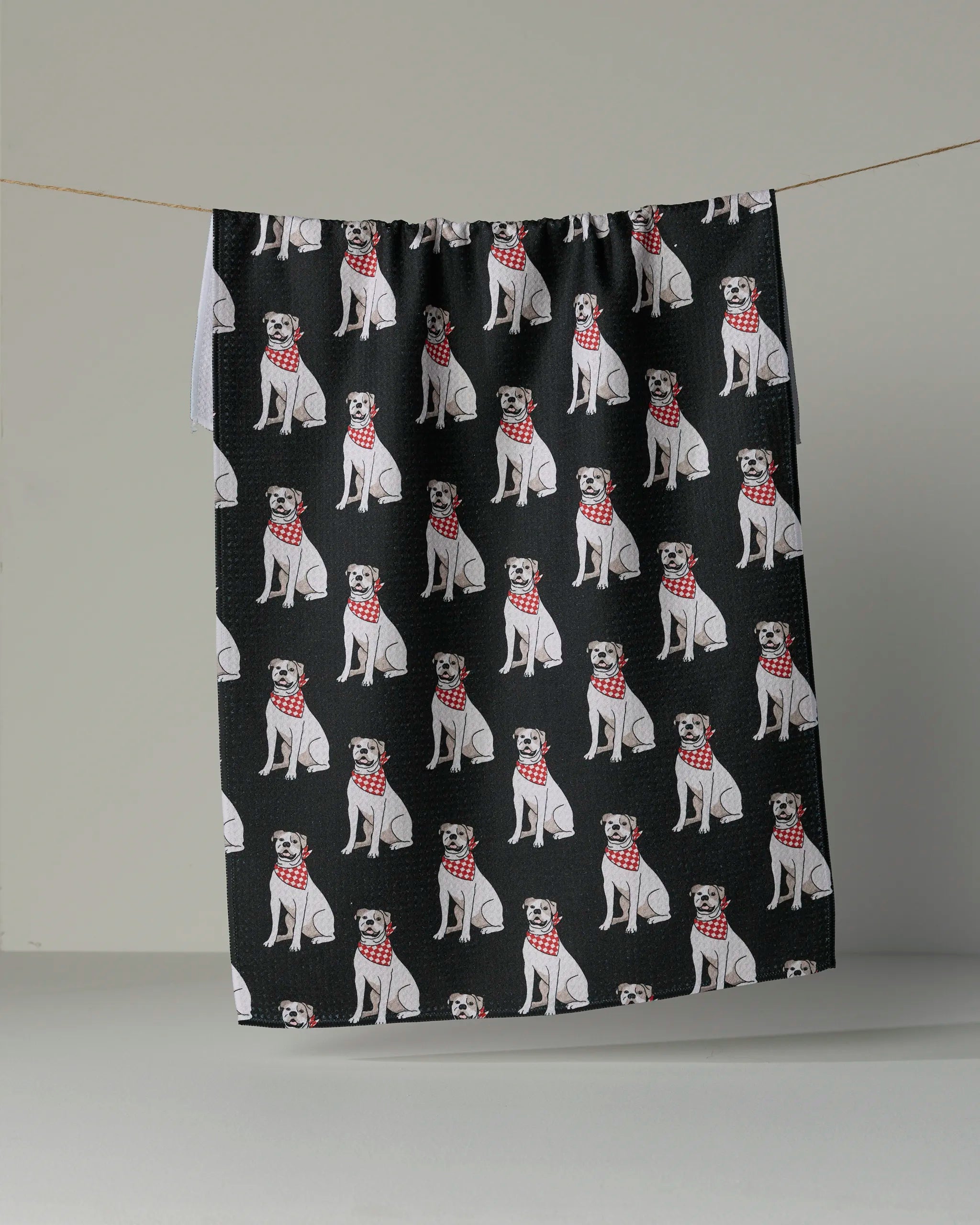 The “Bulldog Club” tea towel by Geometry is made from recycled materials and showcases a repeating pattern of white dogs in red plaid scarves on black fabric, displayed against a plain gray background.