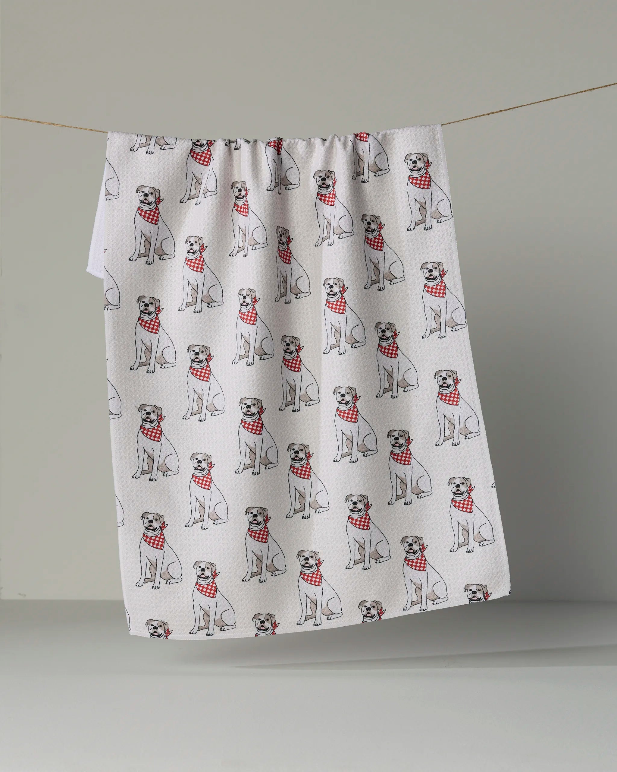 The Geometry “Bulldog Club” tea towel features a repeated pattern of cartoon bulldogs sporting red plaid bandanas on a white background. Made from quick-drying, recycled materials, this charming and sustainable kitchen accessory brings both style and eco-consciousness to your home.