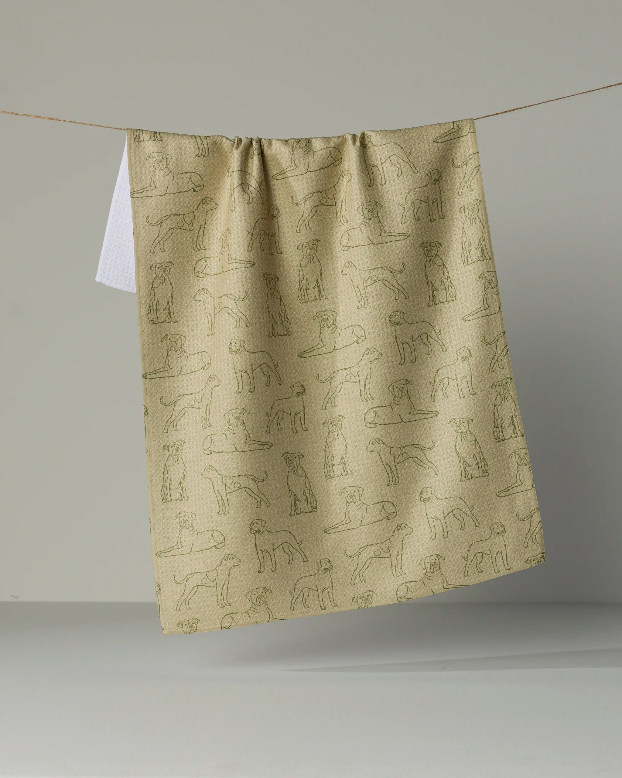 A beige, eco-friendly “Bulldog Pups” kitchen towel from Geometry, featuring line drawings of various dog breeds, hangs on a clothesline against a plain background.