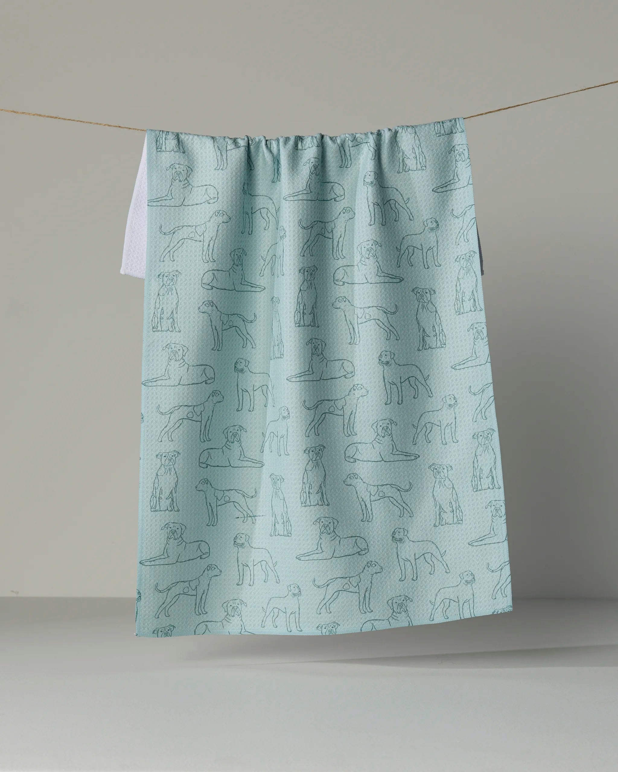 A light blue, eco-friendly “Bulldog Pups” tea towel by Geometry, featuring outlined drawings of various dogs, hangs on a clothesline against a plain, neutral background.