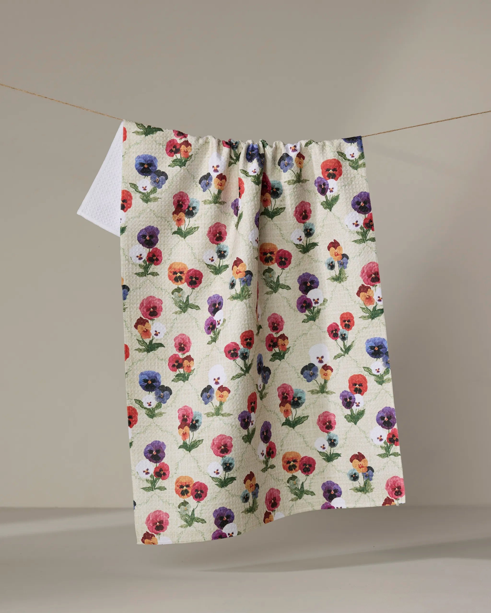 The "Bunch of Pansies" reusable kitchen towel by Pascale Dilger, featuring a colorful pansy flower pattern, hangs on a clothesline against a neutral background.