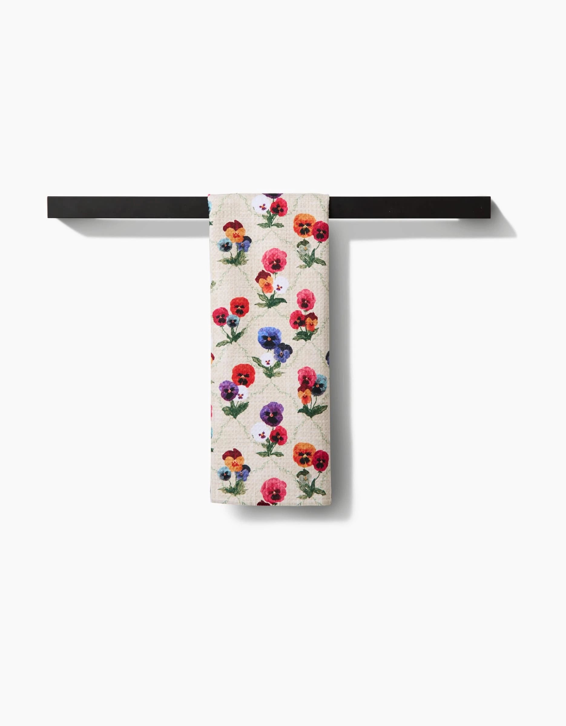 The Pascale Dilger “Bunch of Pansies” tea towel, featuring a vibrant floral design, hangs on a black bar against a white background. Made with Geoweave fabric, it offers exceptional absorbency and durability.