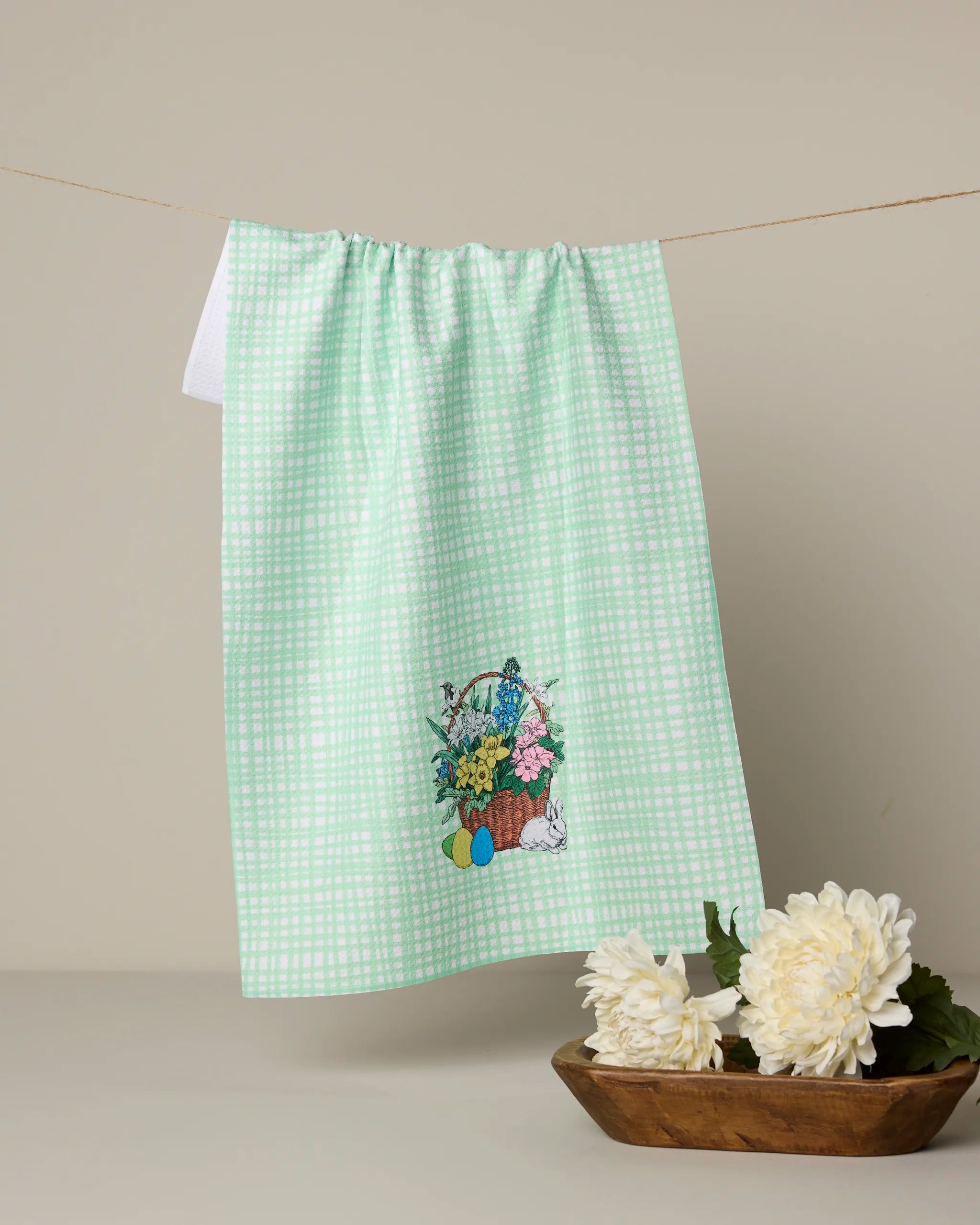 The Geometry Bunny Basket kitchen towel, made from recycled materials, features a green and white checkered pattern with embroidered flowers and Easter eggs. It hangs on a clothesline above white flowers and a wooden bowl.