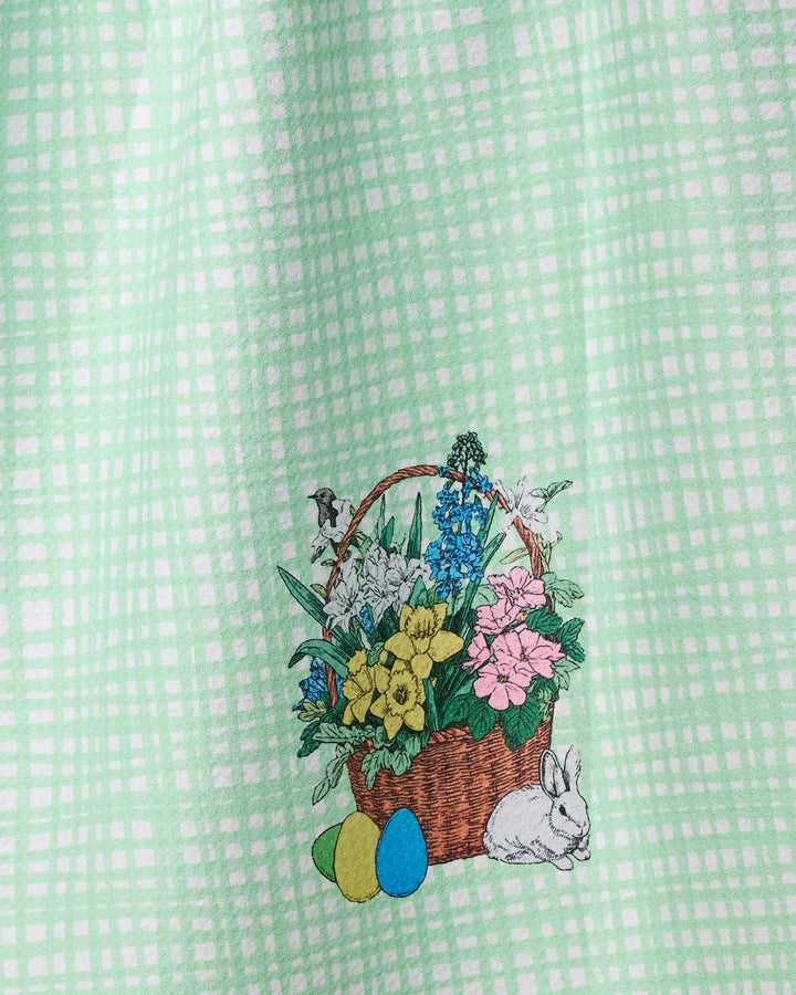 The Geometry Bunny Basket kitchen towel features a green and white plaid design with a woven basket filled with colorful flowers, three painted eggs, a white bunny, and a small bird.